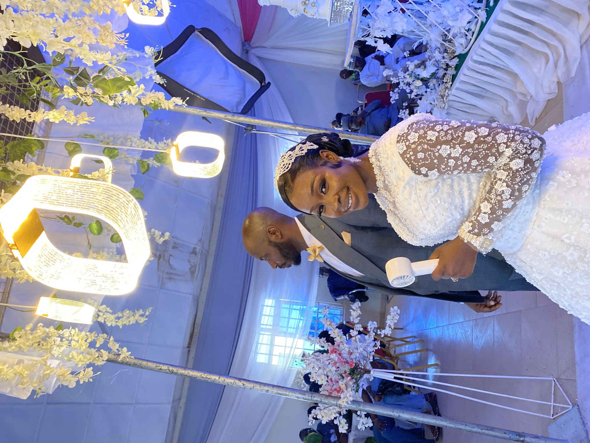 The scene depicts a wedding celebration in Dutsen Kato, Nigeria. A woman in a white wedding dress with lace sleeves and a tiara smiles at the camera while holding a small white fan. A man in a gray suit with a gold boutonniere stands beside her. They stand near a table decorated with white flowers and what appears to be a tiered cake. In the background are seated guests and elements of the venue's decor, including hanging lights and white draped fabric.
