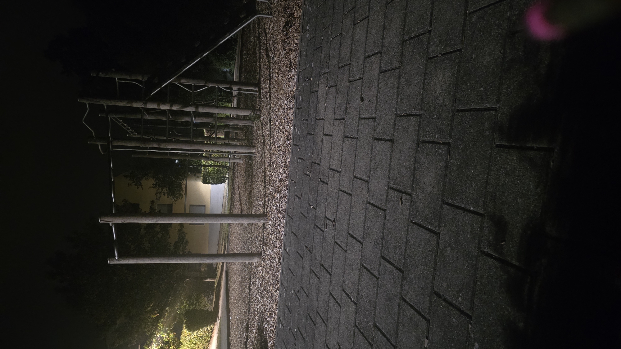 The image captures an outdoor scene at night, likely a park or playground, in Stuttgart, Germany. In the foreground, a dark grey paved pathway made of rectangular interlocking bricks is visible. The pathway is wet, reflecting some ambient light. The bricks are arranged in a regular pattern.
Beyond the pathway, the ground is covered with a layer of dry, brown leaves, creating a textured surface. This area appears to be a soft landing surface for playground equipment.
Dominating the mid-ground is a sturdy wooden playground structure, possibly a climbing frame or monkey bars. It consists of thick, vertical wooden poles supporting horizontal bars and a ladder-like climbing section. To the right, a dark slide descends from the structure. The structure is illuminated by an unseen light source, casting shadows.
In the background, a building with yellow-toned walls and windows can be seen. The building is partially obscured by trees and bushes. Dense, dark green foliage lines the edges of the property. The overall ambiance is quiet and somewhat eerie due to the nighttime setting and the artificial lighting. The sky is dark and overcast, typical for a night in Stuttgart. A small, blurred pink object is visible in the lower right corner of the image, suggesting the presence of a person or an object held by a person just outside the frame.