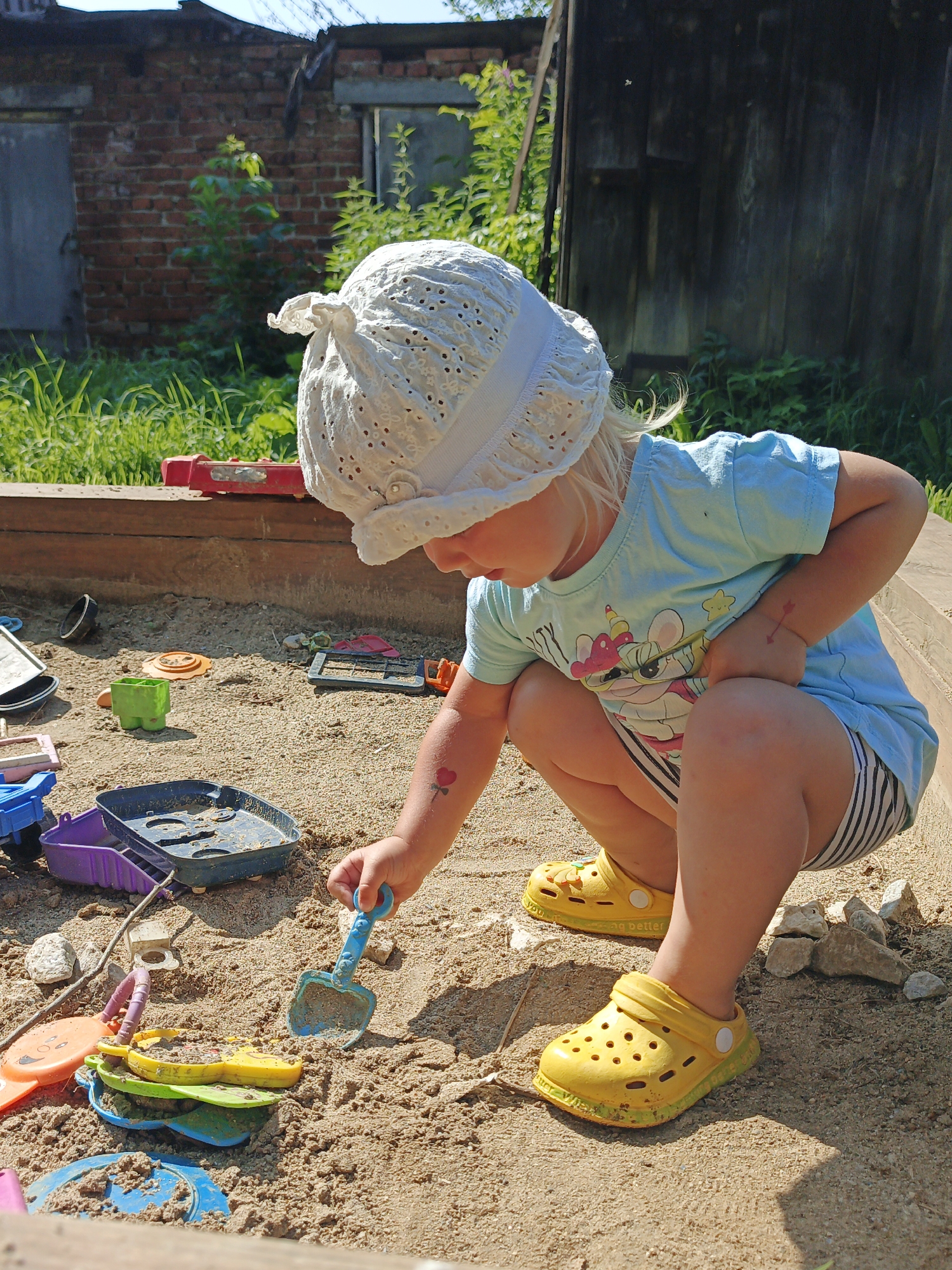 The image shows a young child playing in a sandbox, with a red brick building and a wooden fence in the background, indicating a location in Medvedyovka, Russia. The child, wearing a white hat, a blue t-shirt with a cartoon print, and striped shorts, is squatting and using a blue plastic shovel to play in the sand. Scattered around the sandbox are various plastic toys, including buckets, molds, and a small blue toy truck. The sand is dry and granular, with some small pebbles mixed in. The child has a temporary tattoo of a red arrow on their left forearm. The overall scene depicts a child engaged in outdoor play during daylight hours.