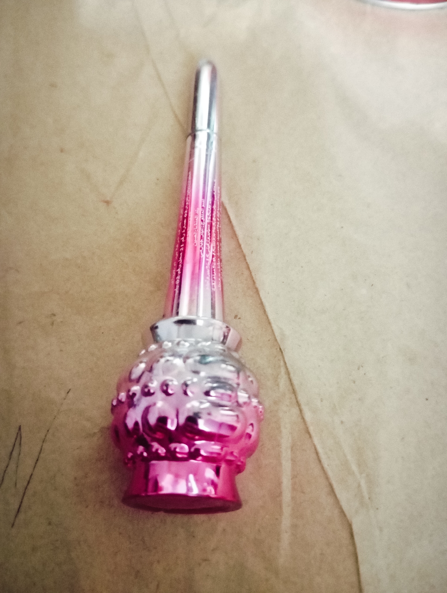 A single, small, pink and silver object resembling a miniature perfume bottle or a decorative Christmas ornament is placed on a textured, light brown surface. The object has a bulbous, textured base with raised circular patterns, and a slender, pointed top that is silver at its very tip, transitioning to pink and silver down its length. The pink portions of the object are glossy and reflective. There appears to be some faint text in a lighter pink, vertically aligned on the pink section of the object's upper part. The background is a flat, slightly wrinkled surface, possibly paper or fabric, with some subtle variations in tone and texture. The lighting is even, casting no distinct shadows, suggesting an indoor setting or diffused natural light. There are no people or other objects visible in the frame. The scene is static, with the object simply being presented on the surface.