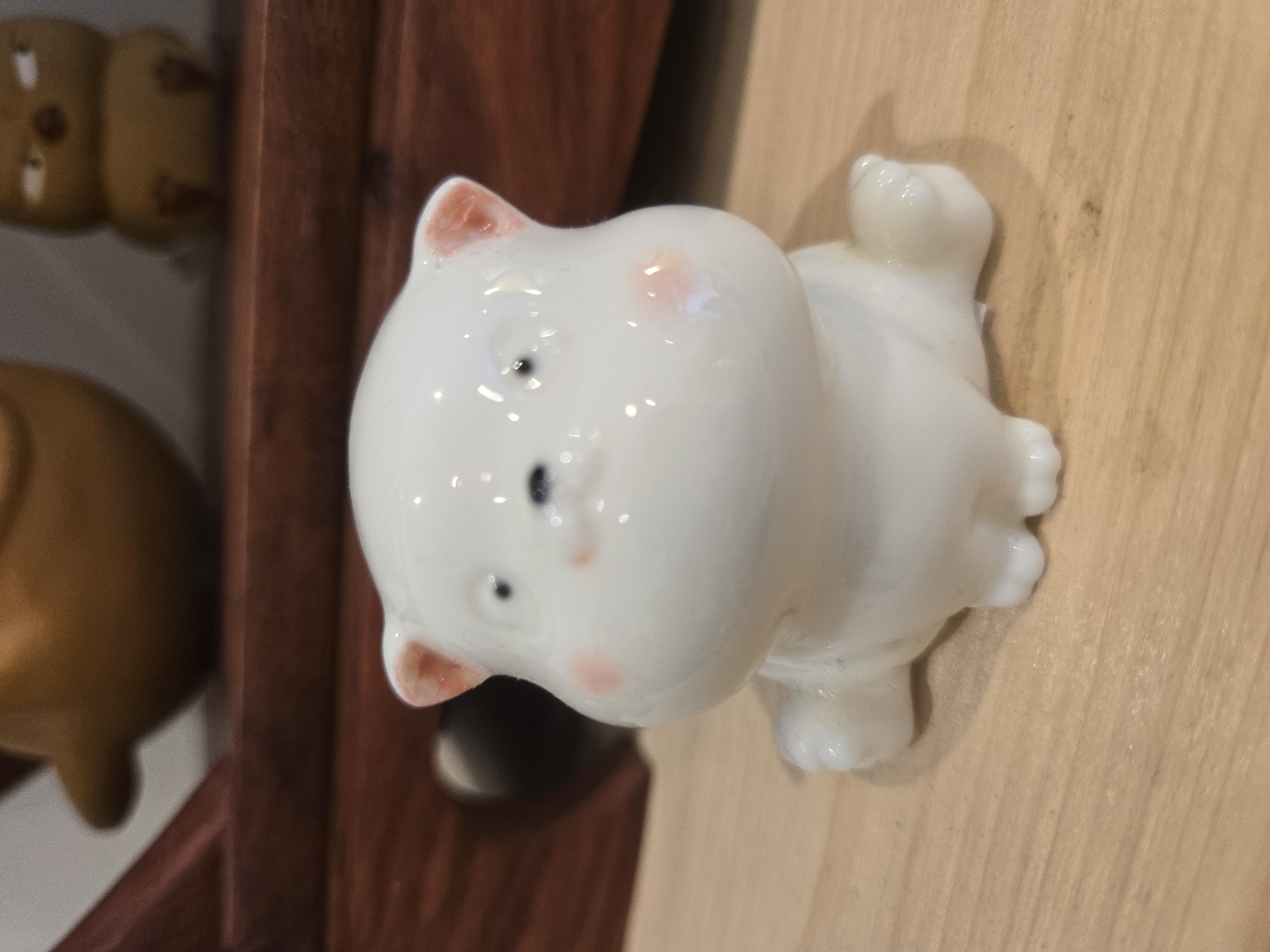The image depicts a small, glossy, white ceramic cat figurine with pink detailing on its ears and cheeks, lying on a light brown wooden surface. The cat is positioned on its side, with its paws and tail visible. In the blurred background, on the left side of the image, there's a dark wood frame and a golden-colored object. The exact location in Budva, Montenegro, cannot be determined from the image.