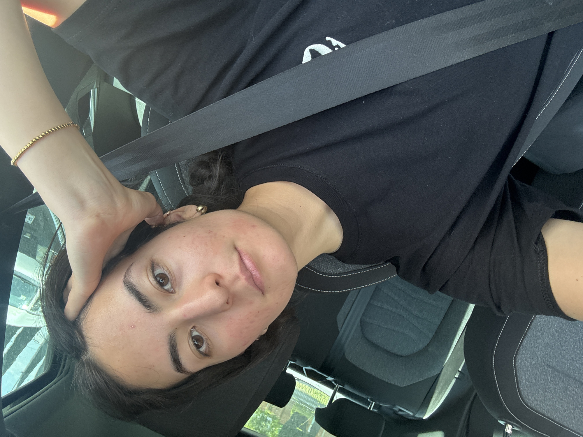 A woman is seated in a car, wearing a black T-shirt and a seatbelt. Her left hand is resting on her head. She has brown eyes and dark hair. A gold bracelet is visible on her left wrist. The car's interior includes grey and black seats with white stitching. The location is unspecified and no evidence suggests it is San Bernardino Tlaxcalancingo, Mexico.