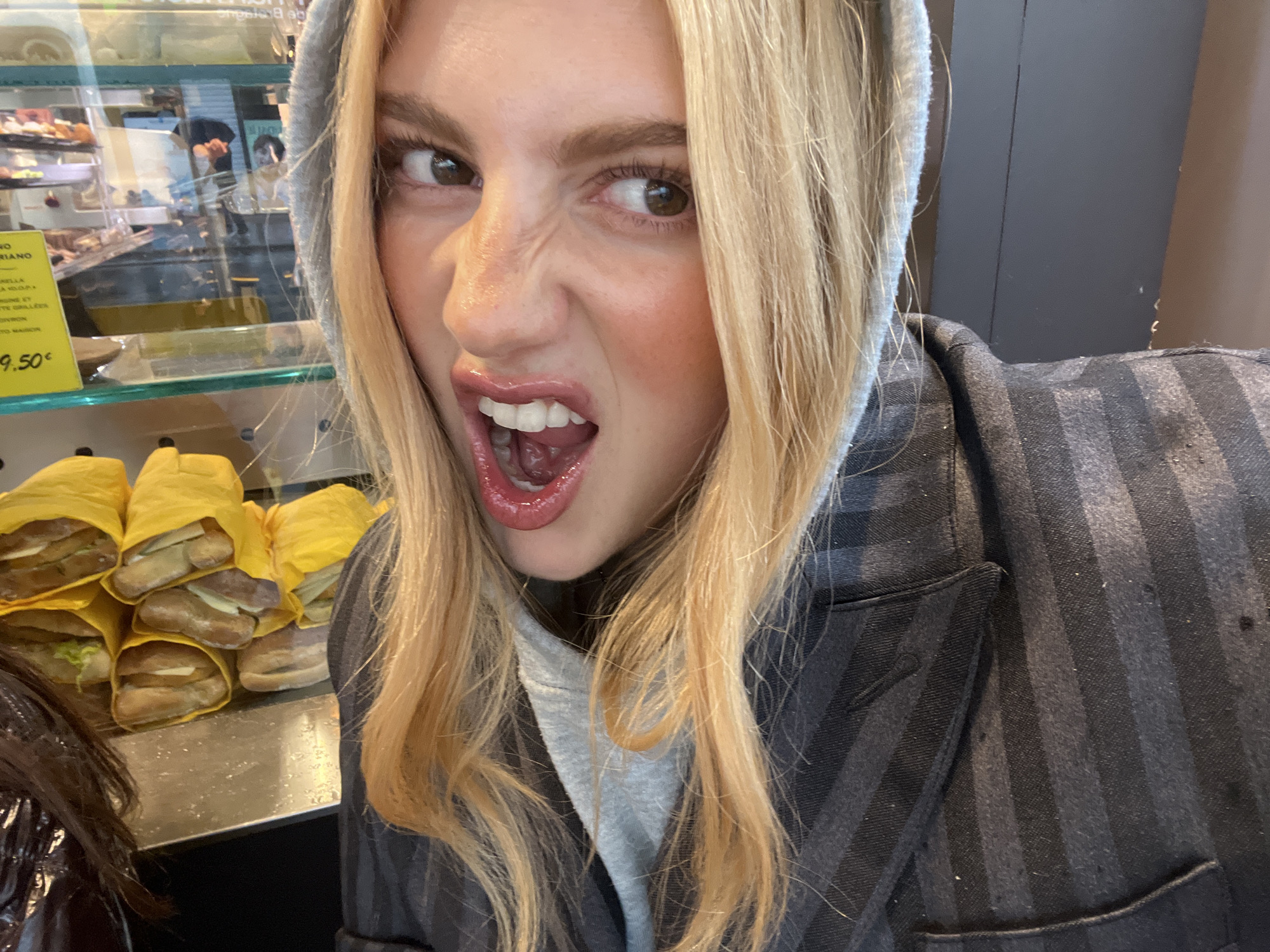 A young woman with blond hair and wearing a gray hooded sweatshirt underneath a gray striped jacket is taking a selfie. She is making a playful, exaggerated facial expression with scrunched-up nose and open mouth. Behind her, to her left, is a glass display case filled with pastries and sandwiches wrapped in yellow paper. A sign in the display case indicates the price of something is €9.50. Through the glass, reflections suggest the location is in Paris, France, with a shop employee visible. To the bottom left is a partial view of someone wearing a dark jacket.
