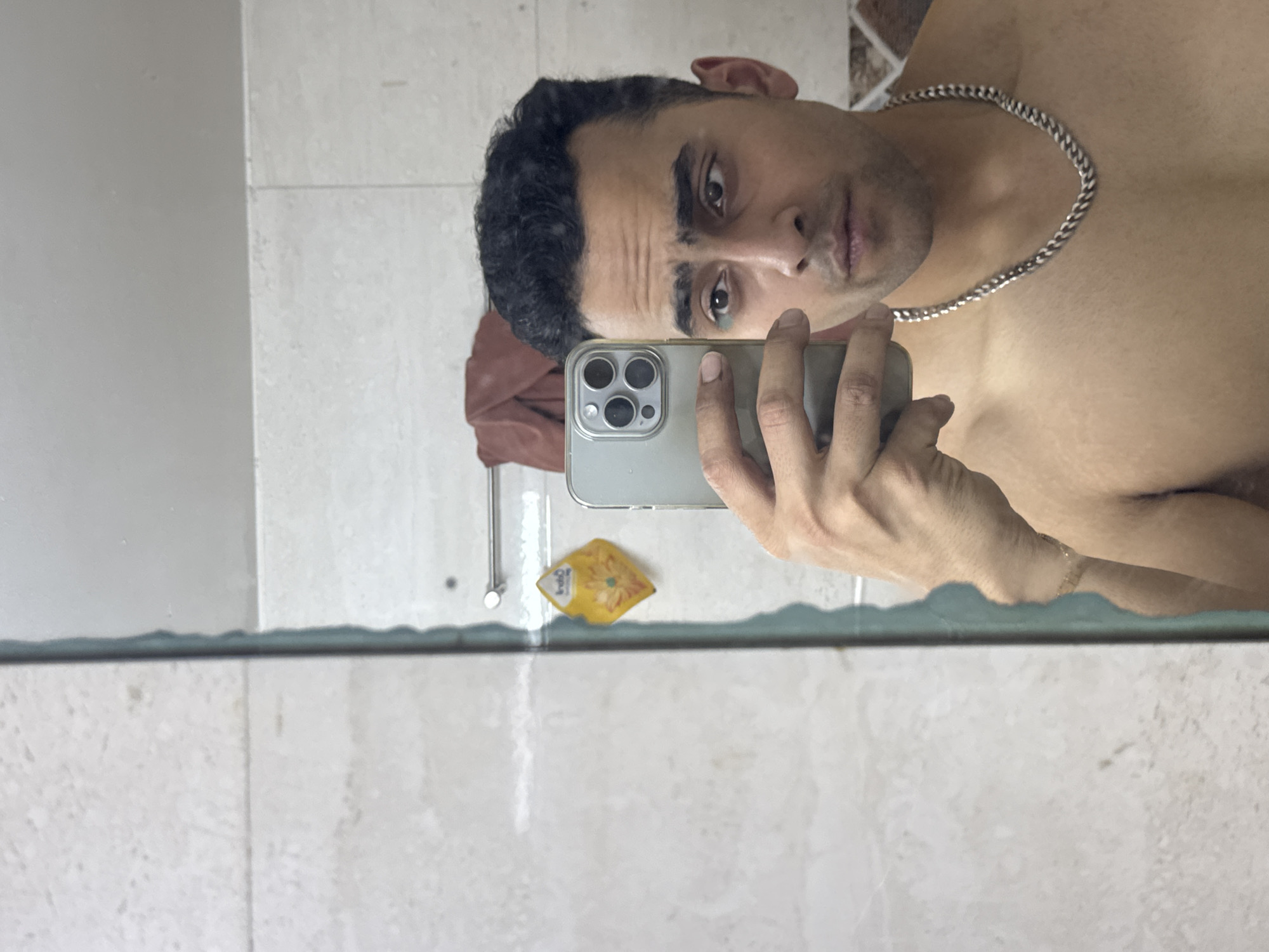 A man is taking a selfie in front of a bathroom mirror. He is shirtless and wearing a silver chain. He is holding a silver iPhone in his right hand. The background includes white tiled walls. A yellow promotional advertisement for sunflower oil is affixed to the mirror. A red cloth is hung on a hook. It is unlikely that the precise location in Kolkata, India can be determined from the available imagery.