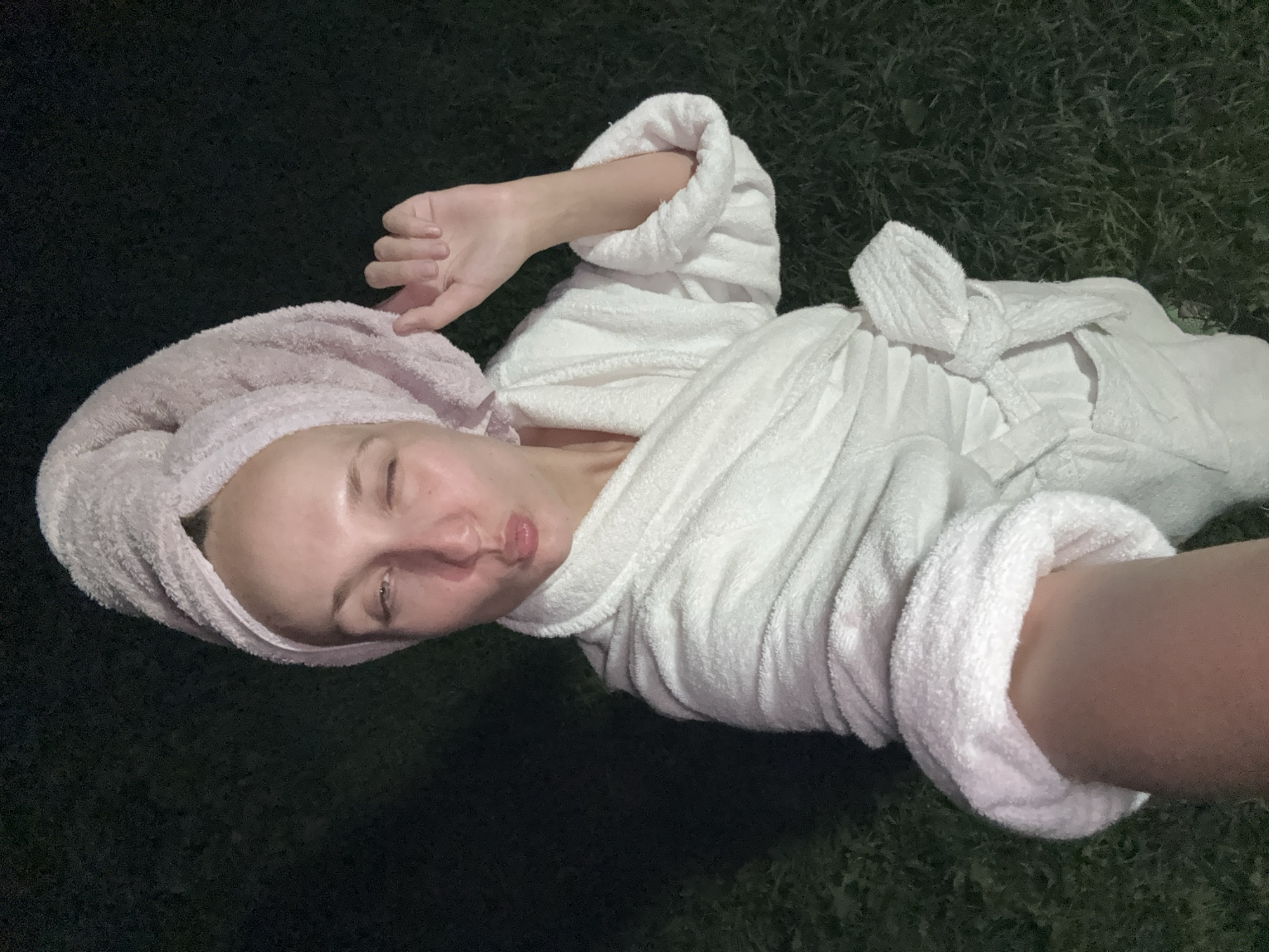 The image shows a woman wearing a white bathrobe and a pink towel wrapped around her head. She is standing outdoors at night, in what appears to be a grassy area. The woman is looking at the camera and making a duck face, with her eyes partially closed. Her right arm is raised, with her fingers splayed. The background is dark and out of focus, but the grass is visible. The image appears to be a selfie taken with a phone. The location is identified as Sharkan, Russia.