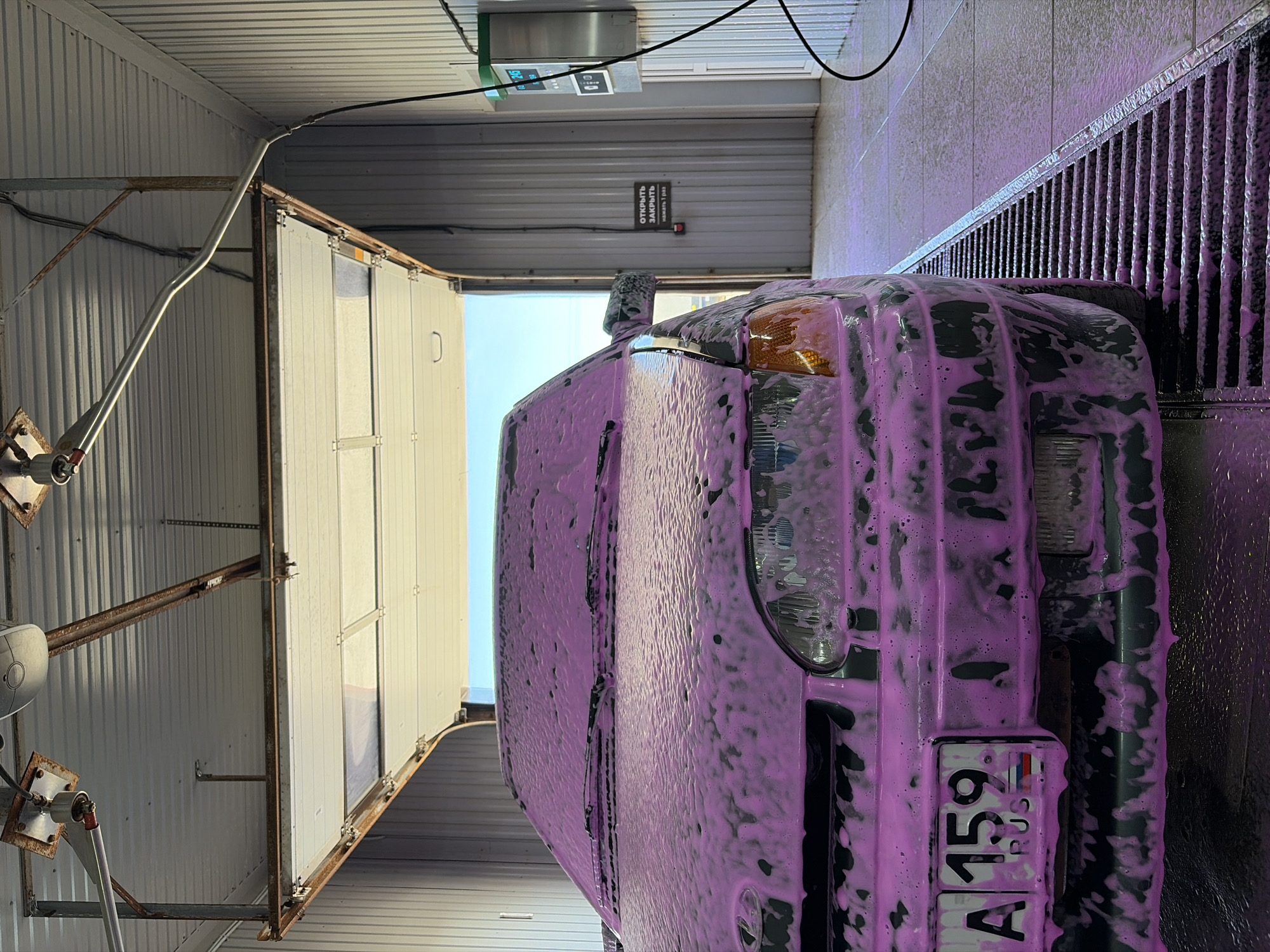 The image depicts a dark-colored vehicle covered in pink foam, seemingly undergoing a car wash process. The car is positioned within a structure with corrugated metal walls and a metal grate visible to the right, suggesting a car wash facility. A control panel is mounted on the wall above the car. The license plate, partially obscured by foam, indicates that the vehicle is from Russia. Based on the license plate's geographical code "59", the location can be inferred to be in the city of Chaikovsky, Perm Krai, Russia.