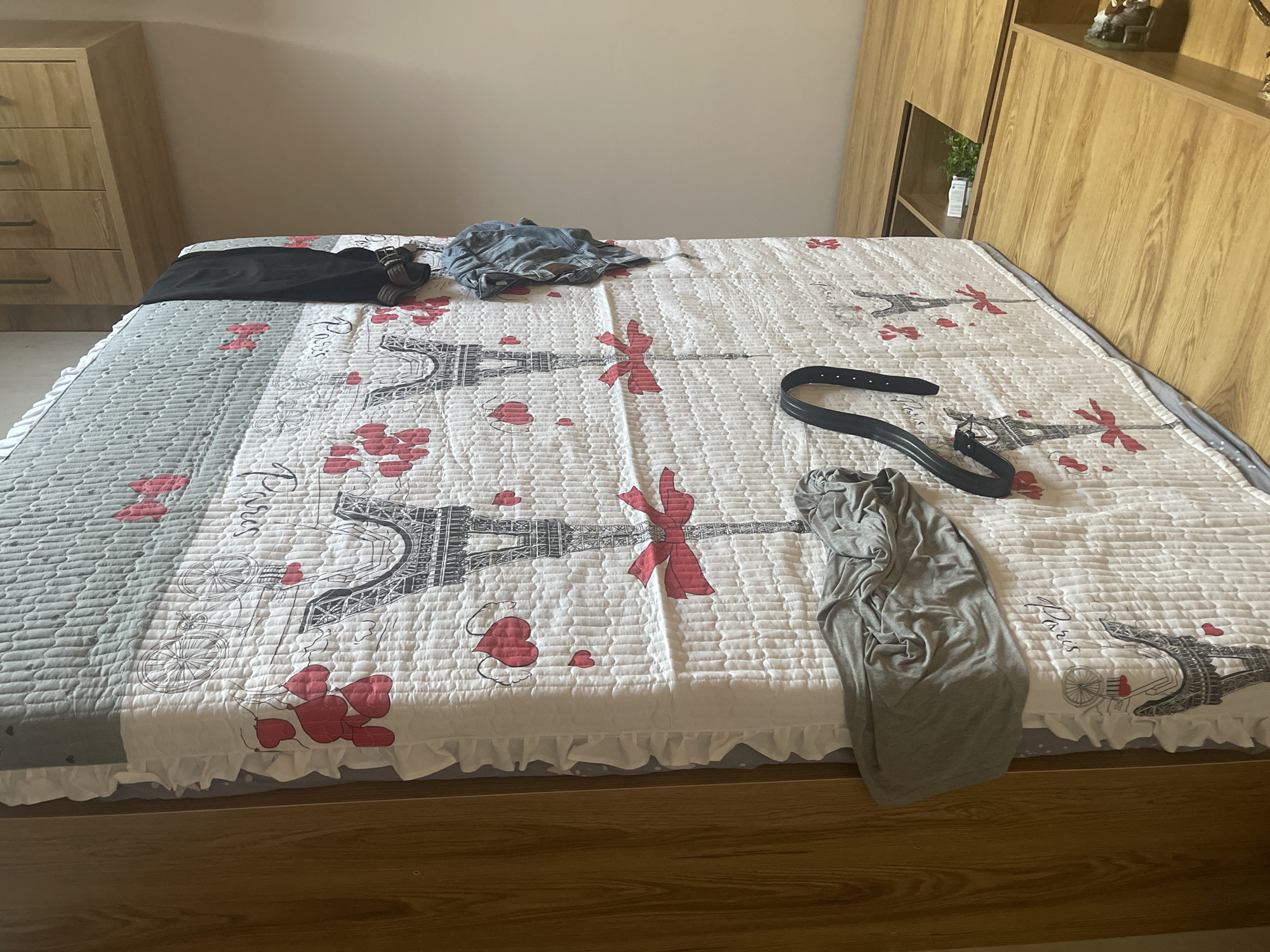 The image shows a messy bed with clothes strewn across the comforter. The comforter is white with a pattern of Eiffel Towers and red hearts. There is a black belt and a grey t-shirt on the bed. To the left of the bed is a light wood dresser with three drawers. To the right of the bed is a light wood headboard with shelves. The overall impression is one of disarray. The image does not provide any specific clues about the location in the city of Qarshi, Uzbekistan.