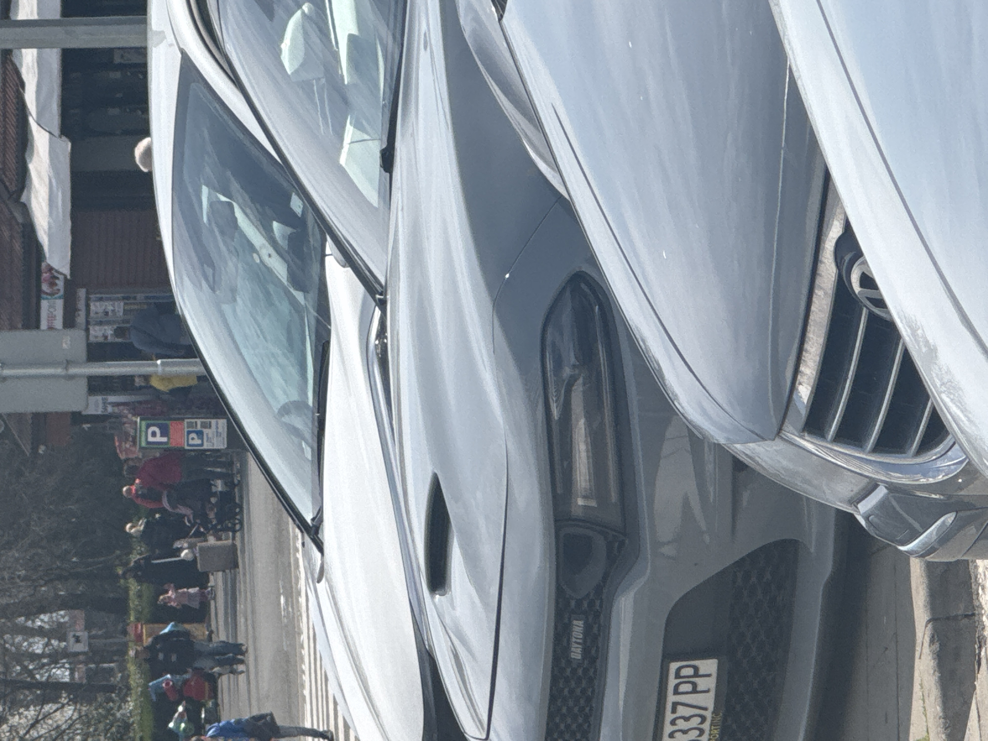 The image captures a street scene likely in Burgas, Bulgaria, inferred from the license plate "337 PP" on the prominently featured vehicle. Several pedestrians are visible in the background, some pushing what appear to be shopping carts. The primary subject is a row of parked vehicles, with the foreground dominated by the front view of a grey car, possibly a Dodge Charger Daytona, recognizable by the "DAYTONA" branding. The vehicle has a European-style license plate. To the left of the vehicles is a parking sign and some greenery. The overall atmosphere is that of a typical city street.