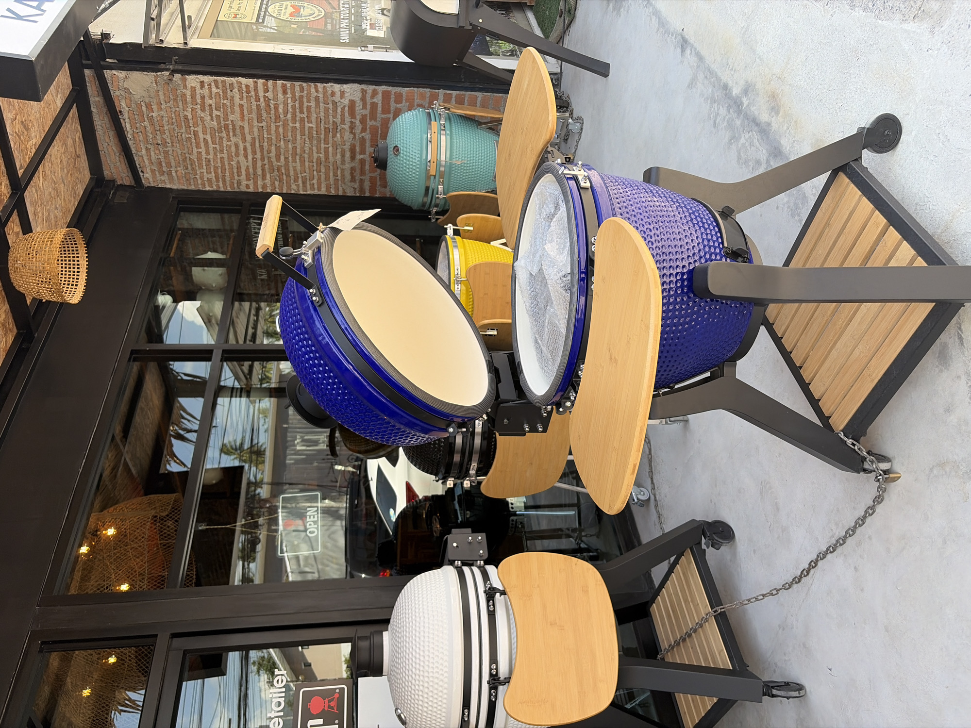 The image captures a storefront in the city of Ko Samui District, Thailand, showcasing several ceramic grills. The storefront features large glass windows, one of which displays an "OPEN" sign. The grills are arranged on a concrete surface, with some linked together by chains and a padlock. The foreground is dominated by a blue ceramic grill with its lid open, revealing its interior. To the left, a white ceramic grill is visible, also with a wooden side table. In the background, other ceramic grills in teal and yellow are present, along with a black outdoor oven. The environment appears to be a commercial area, possibly a showroom or a retail store, with elements of outdoor display. The lighting suggests daytime.
