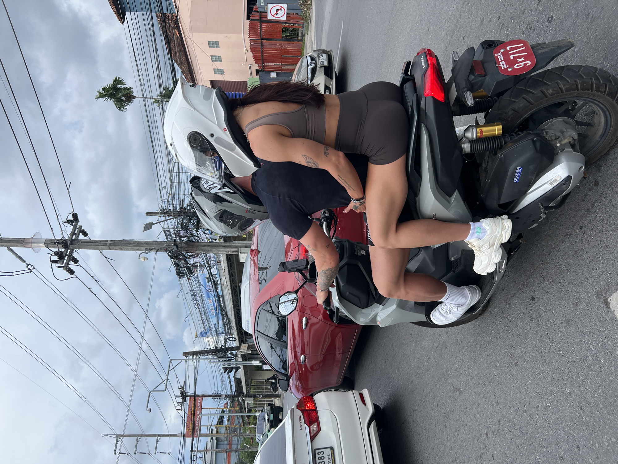 In Chalong, Thailand, two individuals are riding a silver Honda scooter in moderate traffic. The person in front, a male, wears a black t-shirt and a grey helmet. The person behind, a female, has dark hair pulled back and is wearing a brown athletic top and brown shorts. She is wearing white sneakers, and the male is wearing white sneakers. The scooter has a red license plate that reads "9x717" in Thai script. The environment is urban with buildings, traffic lights, utility poles with numerous wires, and other vehicles on the road, including a red car and a white car. The sky is overcast.