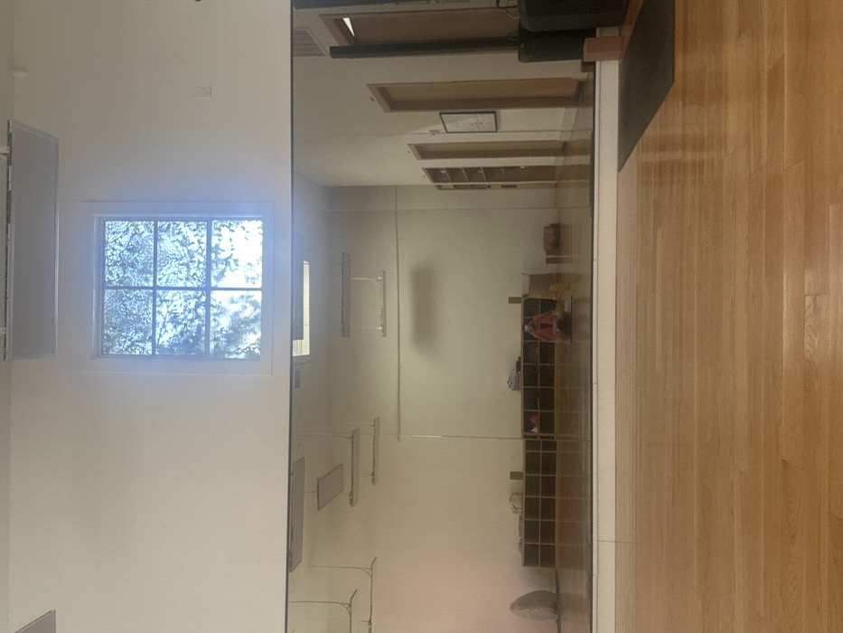The image displays an interior view of a yoga or fitness studio located in Los Angeles, United States of America. The floor consists of polished light-toned hardwood. A large, full-width mirror covers the lower half of the far wall, reflecting the room's interior. Above the mirror, a multi-pane window (two wide, three high) reveals blurred green foliage and bright external light. The ceiling is white, with linear fluorescent light fixtures visible in the mirror's reflection.
Within the mirror's reflection, a low, dark brown wooden cubby storage unit with multiple square compartments is positioned against the far wall. This unit holds various items, including bags and yoga props. To the left of the cubbies, an irregularly shaped, light-colored stone object or sculpture rests on the floor. To the right, a hallway extends, featuring several light-colored wooden doors and a framed picture on the wall.
A single individual, presumed to be the photographer, is visible in the mirror's reflection. This person is seated or kneeling on the hardwood floor near the cubby unit, facing away from the camera, and is wearing a red top. On the actual floor in the foreground, a section of a dark yoga mat is visible extending from the right side of the frame.