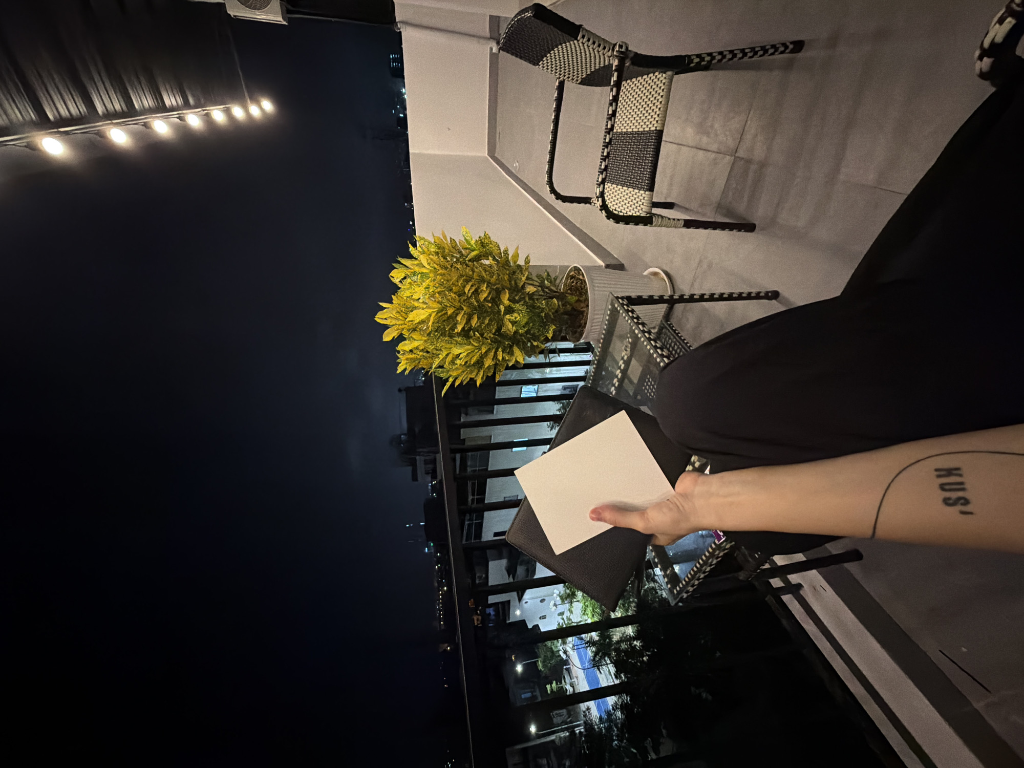 Captured in a high-angle shot, the photograph depicts a nighttime balcony scene in Đà Nẵng, Vietnam. An individual is seated, dressed in black, holding a white square card above a black object in their left hand. The person's right forearm is visible, featuring a tattoo of the word "KUS". A plant in a white pot sits on a small table next to the person. A woven chair is nearby. The balcony overlooks a cityscape illuminated by scattered lights. Above, string lights hang.