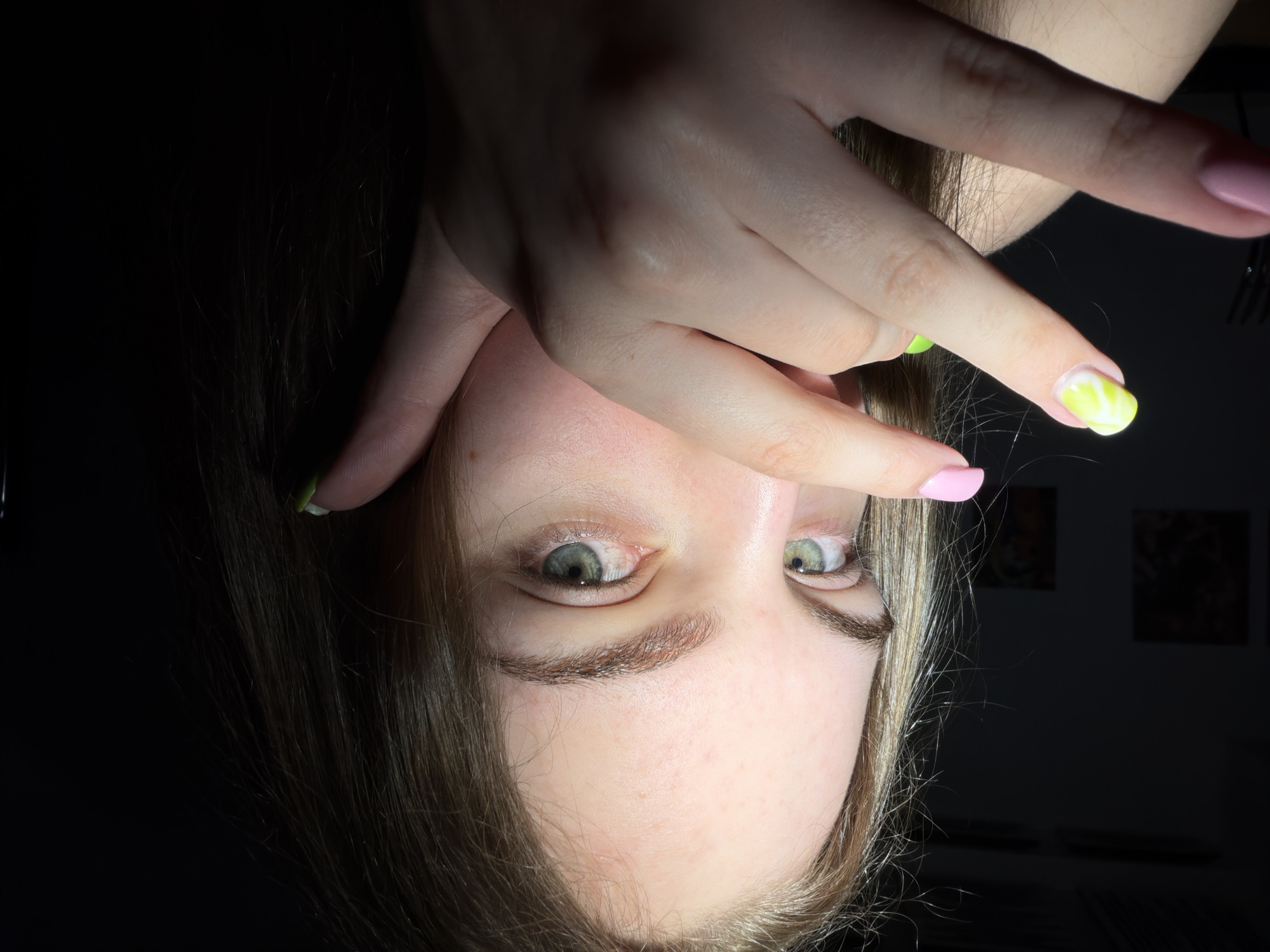 The image shows a close-up, eye-level shot of a young woman with light brown hair and green eyes. She is partially obscuring her face with her hands, which have light pink and lime green nail polish. The background is dark and out of focus, with what appear to be two framed pictures on a wall to the left. The lighting is dramatic, with a spotlight creating strong shadows. No specific location in Dnipro, Ukraine is visible or identifiable.