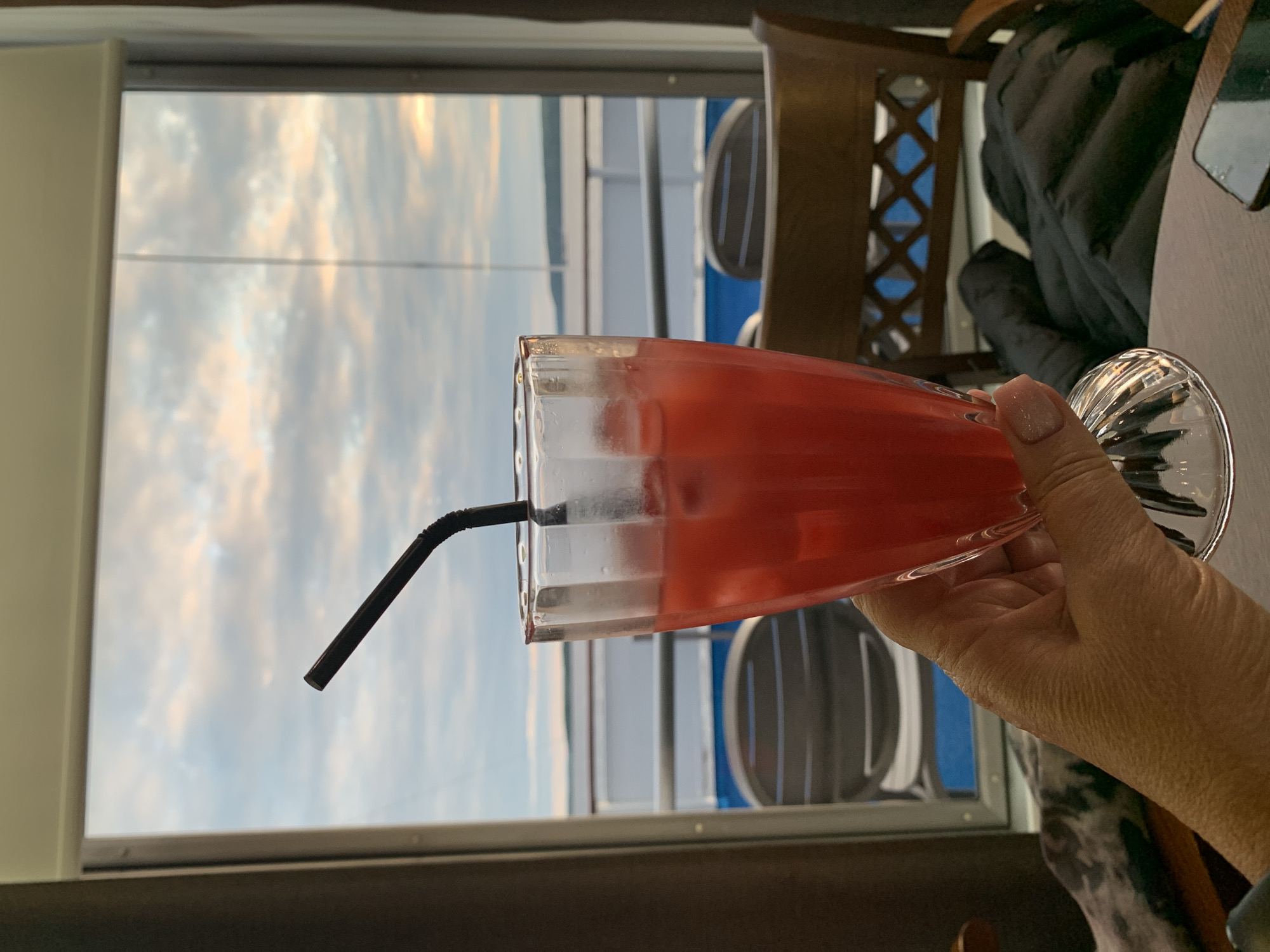A hand holds a glass filled with a red beverage and ice, with a black straw in it. The glass is positioned in the foreground, obscuring part of a window. Through the window, a cloudy sky and a body of water with a white railing and a hint of land in the distance are visible. In the background, chairs and a table are present. The scene appears to be indoors with a view of an outdoor landscape. The location in the city of Taman, Russia is not discernible from the image.