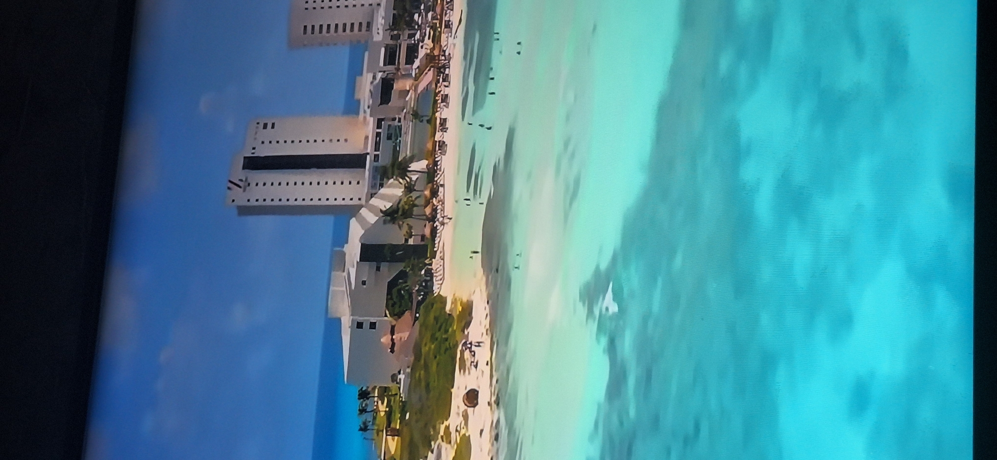 The image shows a wide shot of a tropical resort with a beach and ocean. There are several multi-story hotel buildings in the background, with a distinctive "V" logo on the tallest one. The beach has thatched-roof umbrellas and lounge chairs, and several people are visible walking on the sand and swimming in the clear turquoise water. The ocean extends to the horizon under a blue sky with scattered clouds. There are palm trees on the property. The image is a vertical shot taken from an aerial perspective. The location is not Bila Tserkva, Ukraine, as the scenery is clearly a coastal resort in a warm climate.