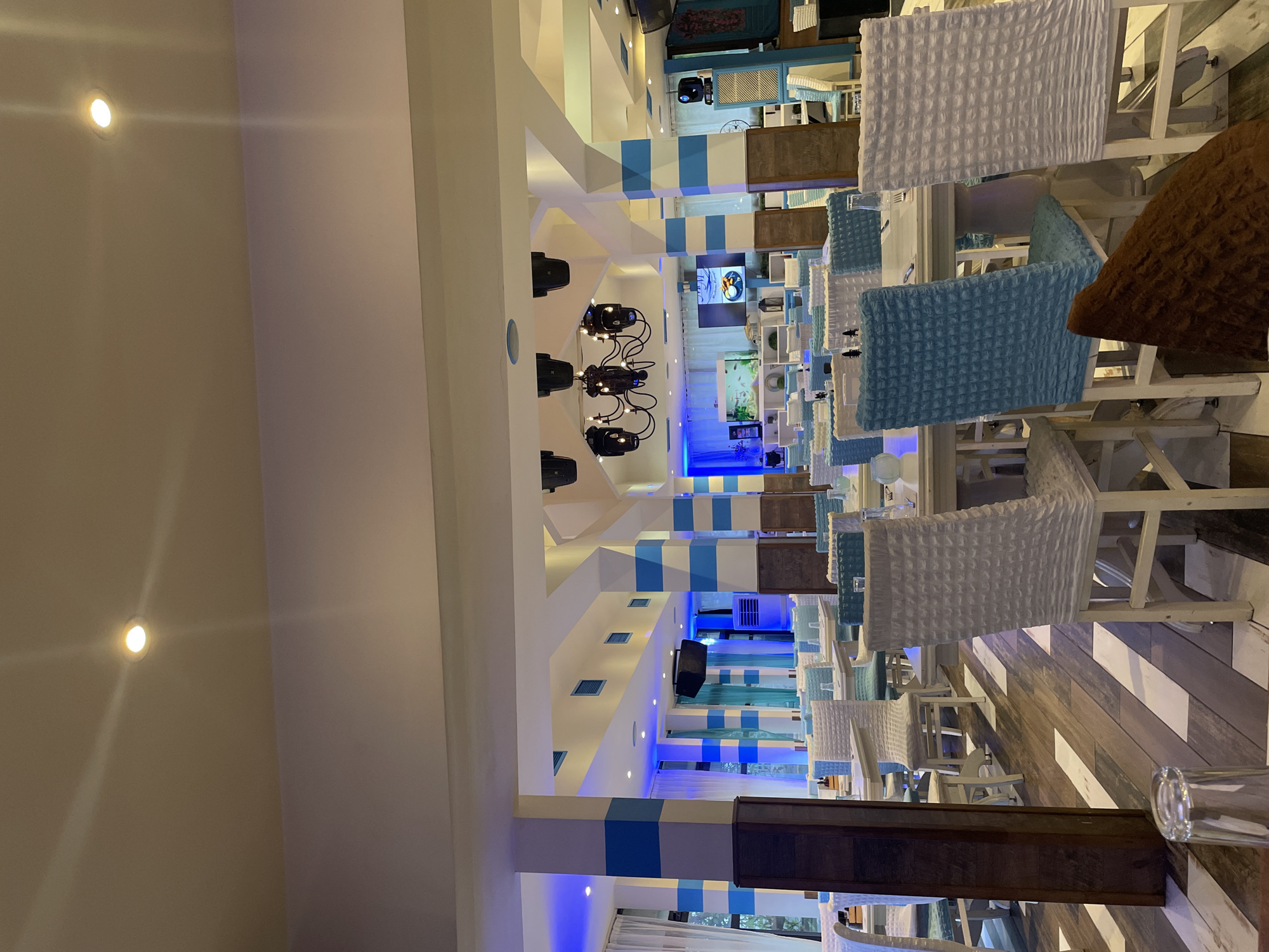 The image captures the interior of a brightly lit, modern restaurant or bar, possibly at night or in the evening, given the reliance on artificial lighting. The scene is devoid of people, presenting a ready-to-use dining space.
The dominant color scheme is white and various shades of blue, giving the venue a clean, possibly nautical or coastal aesthetic. Numerous high-back chairs are arranged around tables, featuring textured covers in white and light blue, with some chairs displaying brown covers. The tables are predominantly white, suggesting a fresh and simple design.
Architectural elements like pillars and wall sections are adorned with alternating white and blue stripes. The ceiling features recessed spotlights and ornate dark-colored chandeliers with multiple light sources. Blue LED strip lighting accentuates certain architectural lines, casting a cool, ambient glow throughout the space. The floor is notable for its diagonal pattern of alternating light and dark wood-like planks.
In the background, an aquarium with fish is visible, adding to the decorative elements. Several display screens are mounted on walls, showing various images, including what appears to be a food item on one screen. Wall-mounted speakers suggest the provision of background music or sound. A clear drinking glass is visible in the foreground on the right. The overall impression is one of a contemporary and inviting dining environment. No discernible text is visible on any objects or screens.