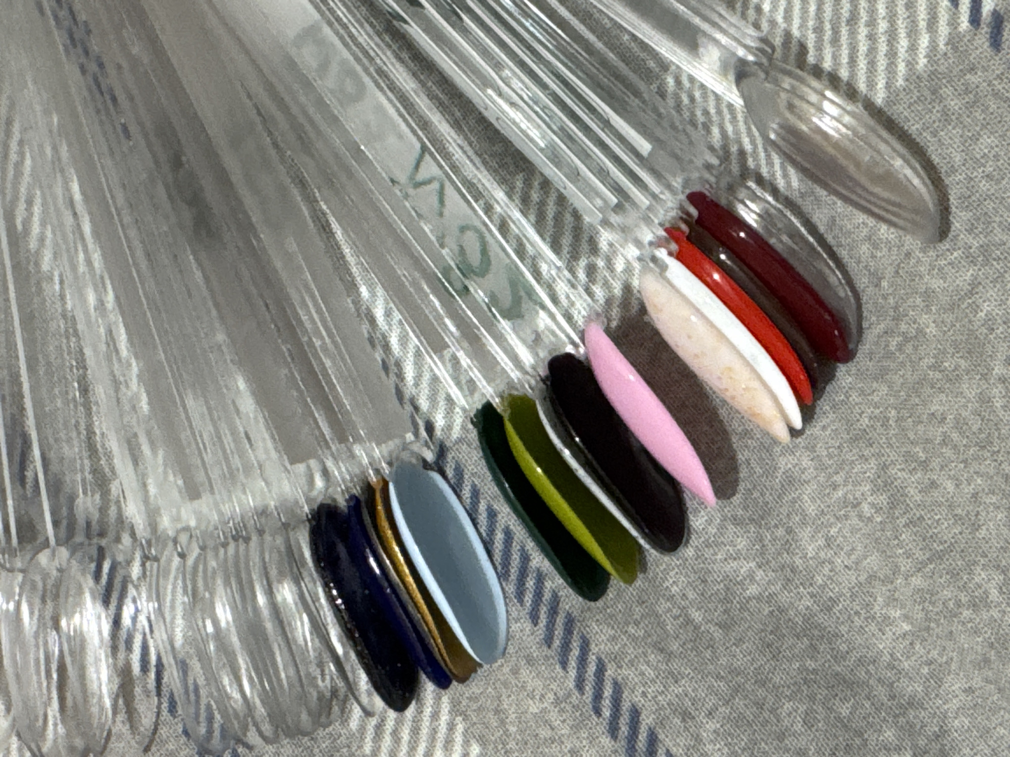 The image shows a set of clear plastic nail polish swatch sticks arranged on a grey textured surface. The swatch sticks are fanned out and each displays a different color of nail polish. From top to bottom and left to right, the visible colors include dark blue, gold, light blue, brown, dark green, olive green, black, pink, a speckled light color, white, red, and dark red. The lighting is overhead and creates highlights on the glossy nail polish colors. The background surface appears to be a fabric, possibly a blanket or textile. The scene is indoors. There is no indication of the city of Яромінскі сельскі Савет, Belarus, or any specific location within it.