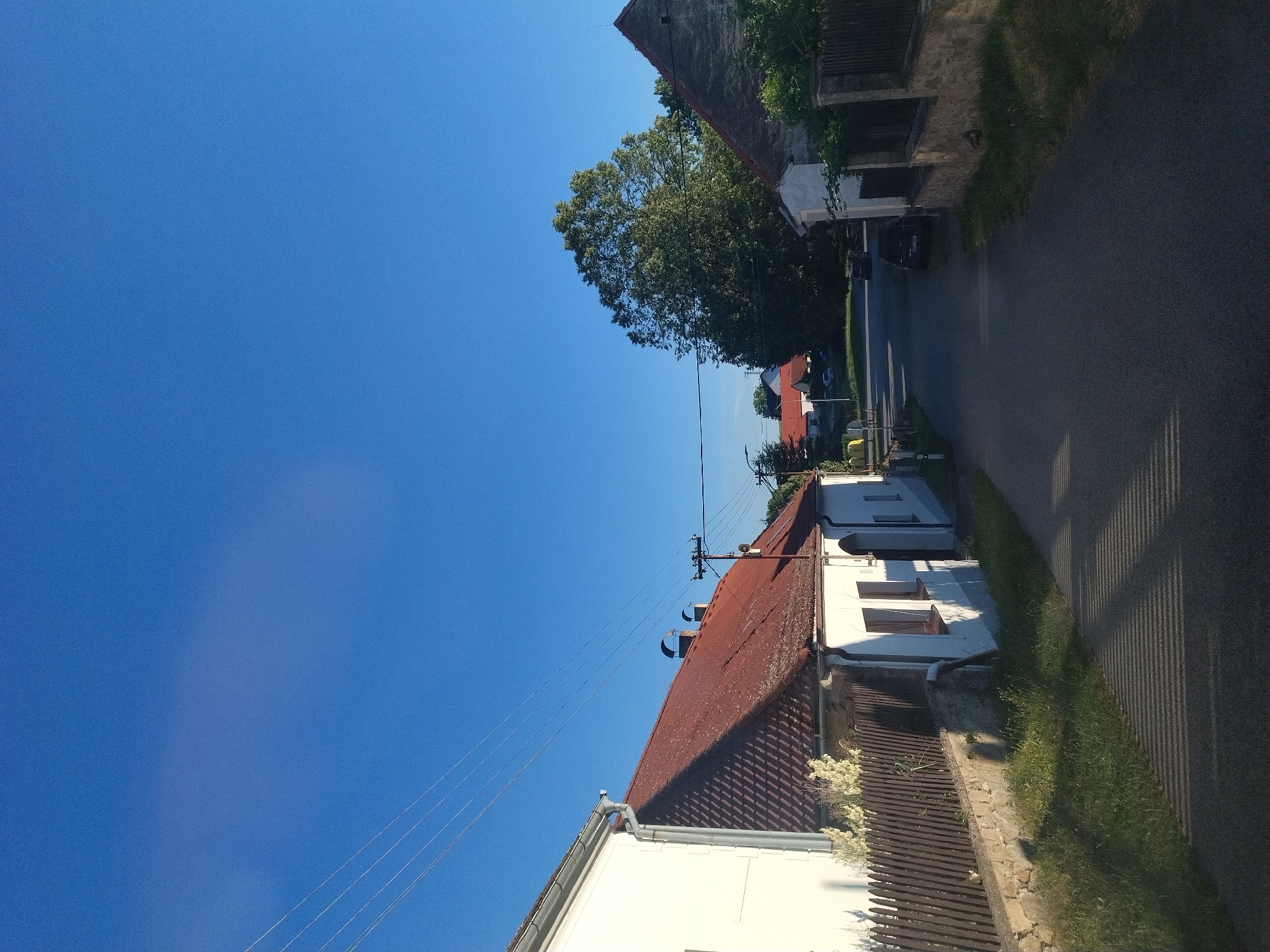The image shows a rural street in Bošovice, Czechia, under a clear blue sky. On the left side of the street, a white house with a red tiled roof is visible, bordered by a wooden fence and a low stone wall. The house has several windows and a prominent gutter. Adjacent to it, a stone wall topped with a wooden fence runs along the side of the street, with a patch of grass and a shadow of striped pattern cast on the asphalt road.
The road itself is paved and appears to be a residential street, with a few parked cars further down. On the right side of the street, another house with a stone foundation and a red tiled roof is partially visible, along with a dark fence. In the background, a large, lush green tree dominates the landscape, with other houses and trees scattered further in the distance. Power lines stretch across the upper portion of the image. The overall scene depicts a quiet, sunlit village street.