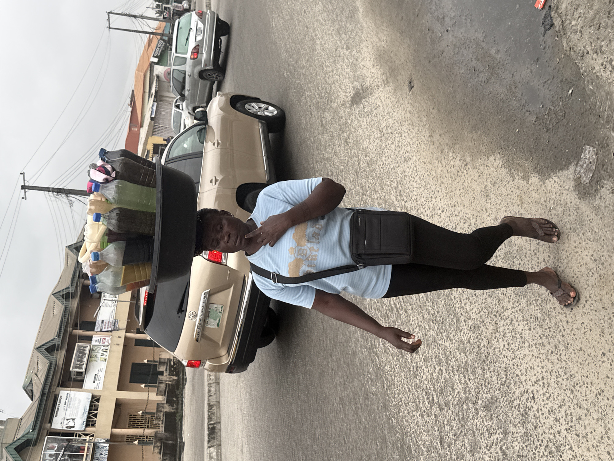 In Ado, Nigeria, a woman is walking on a paved road, carrying a large black basin filled with bottles of liquid on her head. The bottles contain various colored liquids, including yellow, green, and brown. The woman is wearing a light blue t-shirt with a design on the front, black leggings, and brown flip-flops. She has a black crossbody bag slung over her shoulder. Behind her, a gold Toyota Highlander is parked, and a silver SUV is driving in the background. The environment appears to be a street with buildings and utility poles visible. The sky is overcast.