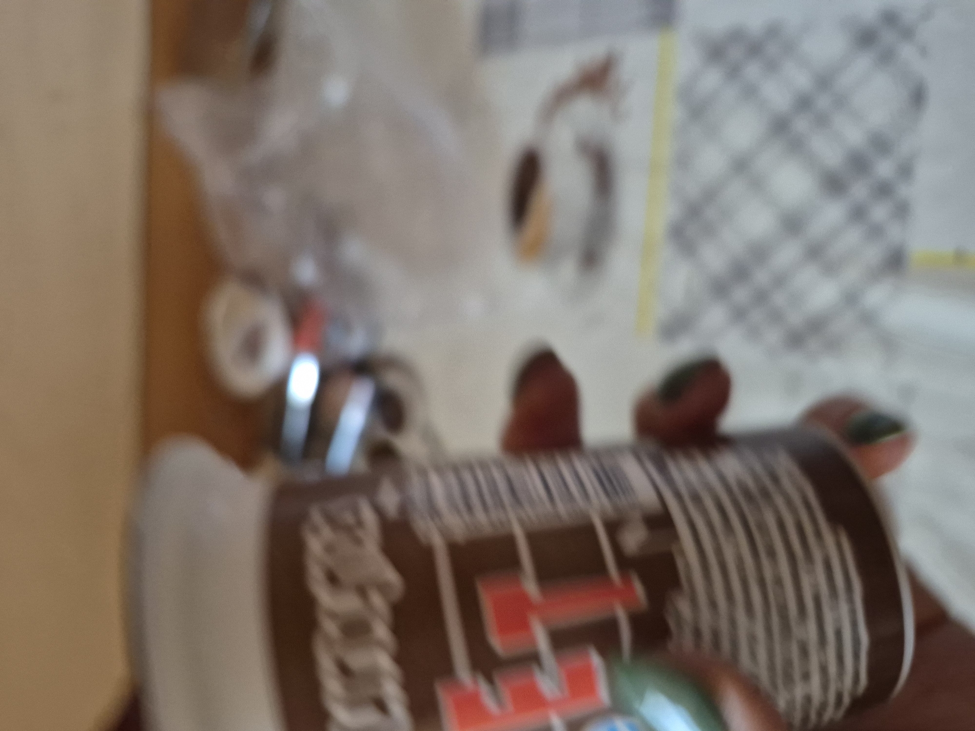 This extremely blurry image, taken indoors, features a human hand holding a cylindrical object, likely a can or bottle. The hand's fingers appear to have dark nail polish, possibly green, on at least one nail visible in the bottom right corner.
The cylindrical object has a dark brown or black label. On this label, prominent red text resembling "E1" is visible, along with indistinct white lettering to its left. A barcode pattern is also discernable on the side of the object.
In the background, to the left, there are blurry shapes that suggest crumpled clear plastic or fabric. Further to the right, a light-colored surface with a distinct checkered or grid pattern is visible, bordered by a yellow strip on its left. Other small, indistinguishable objects are scattered in the mid-ground. The leftmost part of the background shows a plain, light brown wall or surface.
The image captures the simple act of someone holding this object. Due to the severe blur, no specific time of day or emotions can be inferred. There are no visual cues in the image itself to suggest the location is Yerevan, Armenia.