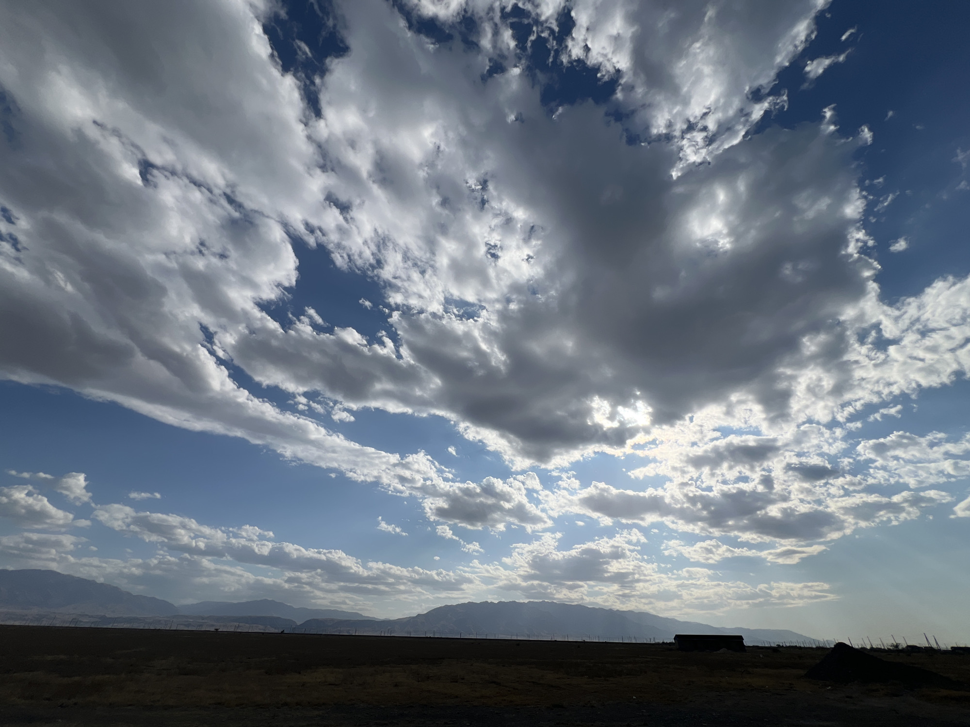 The image depicts a wide-angle view of a landscape under a dramatic sky. The foreground is a dark, barren field with some scattered debris and a few indistinct structures or piles of material in silhouette. A low fence line, consisting of thin vertical posts, runs horizontally across the middle ground. In the distance, a range of hazy mountains stretches across the horizon.
The sky is a dominant feature, filled with a complex array of clouds. Large, heavy, dark gray clouds are interspersed with patches of bright blue sky and lighter, wispy white clouds. Sunlight breaks through in some areas, creating a dappled illumination effect. The overall impression is of an expansive, somewhat desolate natural environment, possibly on the outskirts of a settlement.
While the prompt specifies the location as Ақши, Kazakhstan, there are no specific visual cues within the image that definitively confirm this location. The landscape is generic enough to be found in various arid or semi-arid regions with mountainous horizons.
