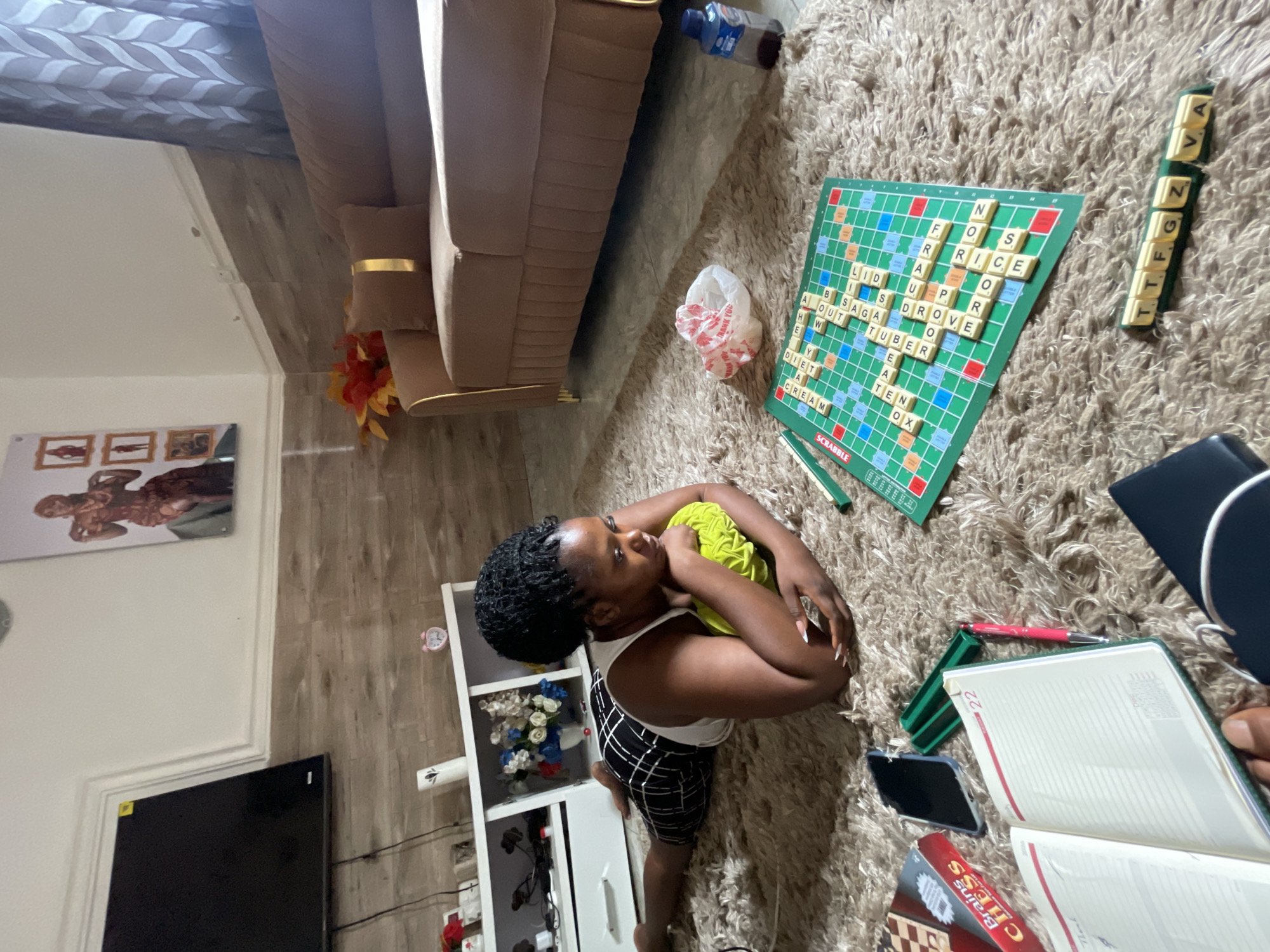 A woman is depicted sitting on a shaggy, light-colored rug in a home interior, deeply engaged in a game of Scrabble. She is positioned on her knees, looking intently at the green Scrabble board laid out on the rug before her. She wears a white tank top and black-and-white patterned shorts, with her hair styled in dark braids.
The Scrabble board is well underway with numerous tiles forming words, including "RICE," "SUPRO," "DROVE," "TUBER," "TEN," "OX," "CREAM," "DIET," "ABOUT," "BSAGA," and "LID." A green tile holder next to the board shows available tiles such as "VA," "TTF," and "GZ." Also on the rug are a white plastic bag with "THANK YOU" printed on it, and a water bottle.
The living space, likely in Kuchingoro, Nigeria, includes a beige sofa with gold trim in the background, next to which artificial foliage is visible. A white shelving unit to the woman's left holds decorative items like flowers and a small pink alarm clock. Further left, a large black television screen is mounted on a wall with wooden-patterned wallpaper. Framed pictures, including one of a muscular figure, adorn another wall. Nearby, an open notebook displaying "22," a red pen, a smartphone, and a box for "BRAINS Chess" suggest other pastimes. The overall scene conveys a relaxed, casual atmosphere, characteristic of leisure time indoors.
