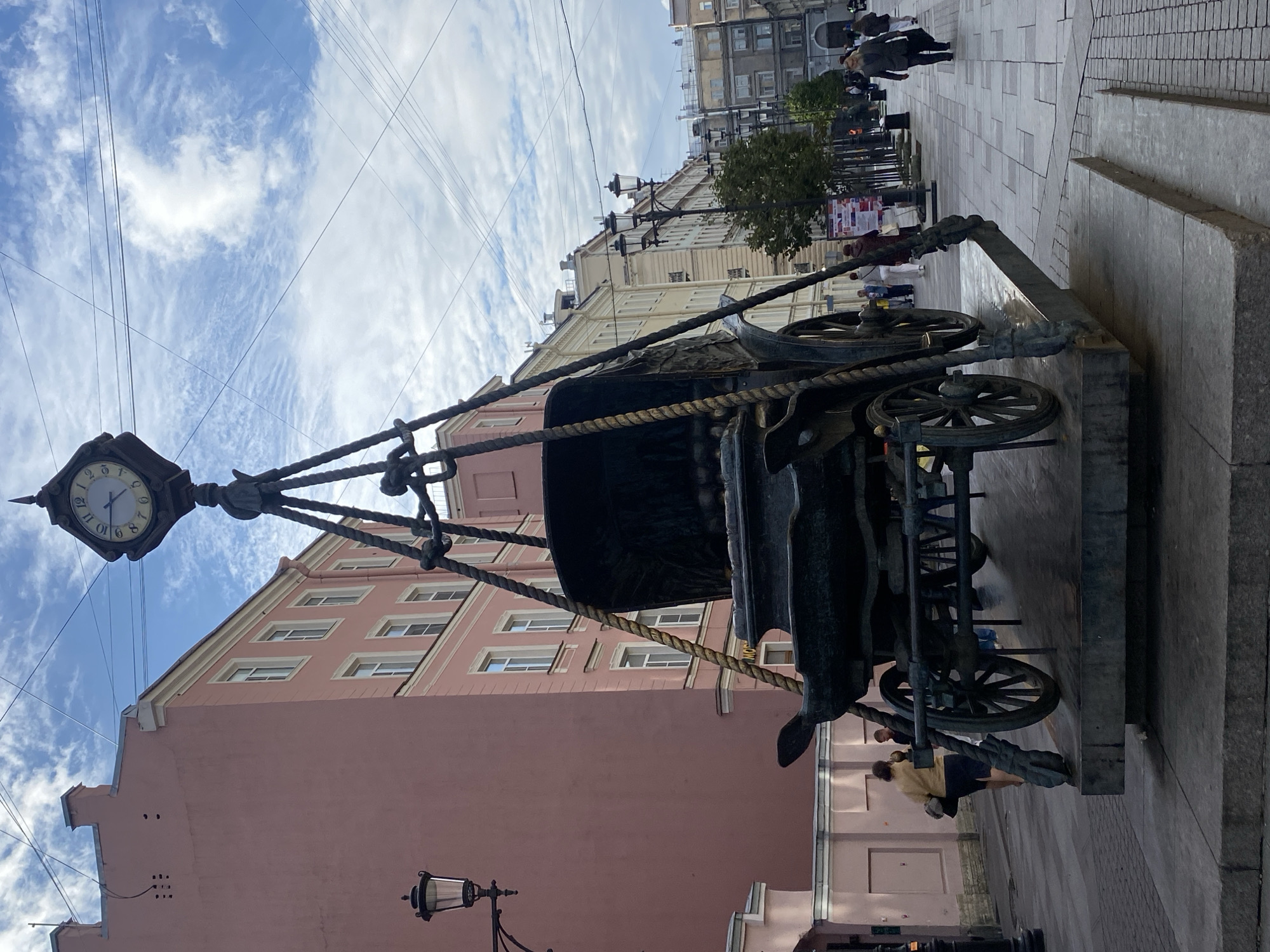 The image depicts a street scene in Saint Petersburg, Russia, featuring a large sculpture of a horse-drawn carriage suspended by ropes from a clock tower. The sculpture is made of dark metal and has intricate detailing. The clock tower itself has a round face with Roman numerals and appears to be functional.
The sculpture is positioned on a paved pedestrian walkway. To the left of the sculpture is a tall, pink building with multiple windows. A lamppost with an ornate design is visible in front of this building. Further down the street, other buildings with classical architecture line the walkway, interspersed with trees and more lampposts.
Several people are present in the scene, walking along the street and sidewalk. They are dressed in casual clothing, suggesting a normal day. The sky is partly cloudy with a mix of blue sky and white clouds. The overall atmosphere suggests a city environment with a blend of historical architecture and public art.
The sculpture is located on Nevsky Prospekt, a main street in Saint Petersburg, near Anichkov Bridge. The clock tower is part of the "Clock and Carriage" sculpture by artist Mikhail Dronov, installed in 2002.