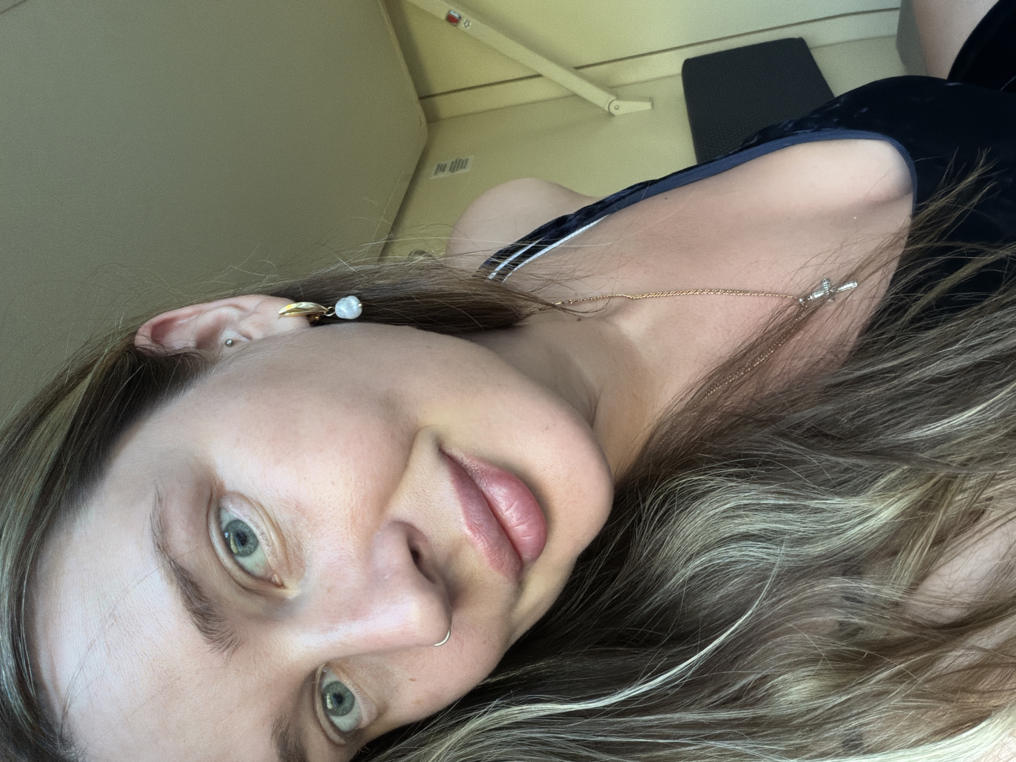 The image shows a close-up view of a woman's face, slightly tilted. She has light skin, light blue eyes, and light brown hair with blonde highlights. She is wearing earrings and a necklace. The background is blurry, but it appears to be the interior of a vehicle, possibly a bus or train, with overhead storage visible. There is no indication of a location in Izhevsk, Russia, or any other specific city.