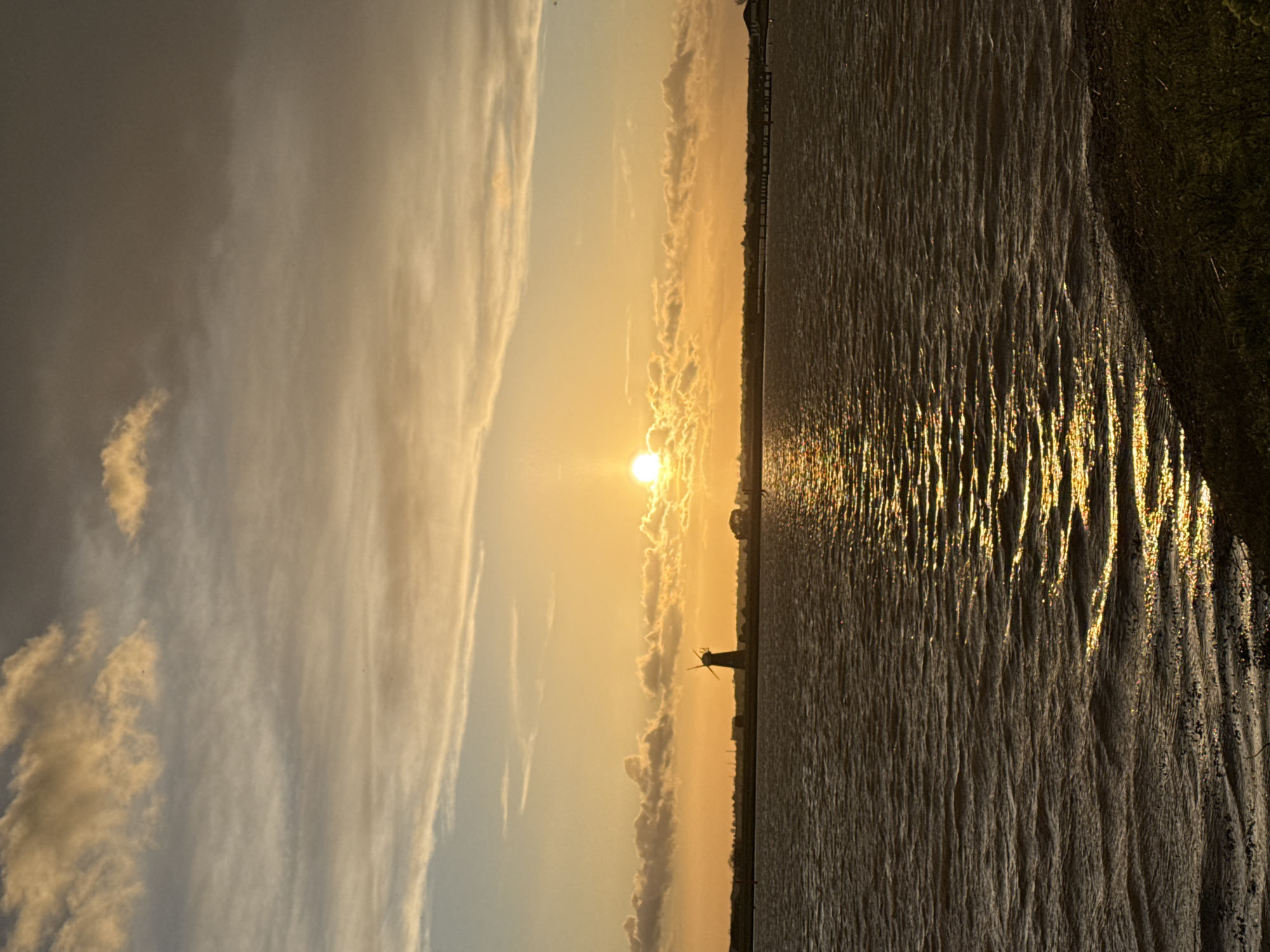 The image displays a sunset scene over a wide body of water, consistent with the Norfolk Broads region near Great Yarmouth, United Kingdom, specifically likely Breydon Water or the River Yare. The sun, a bright golden disc, is positioned low on the horizon, slightly right of the frame's center, partially obscured by illuminated clouds. The sky transitions from vibrant golden-orange near the sun to lighter blue-grey and darker grey-brown in the upper sections, with various scattered clouds. A flat, distant landmass forms the horizon line, featuring the dark silhouette of a four-sailed drainage windmill positioned to the left of the sun. Further along the horizon, other faint structures, including a building with a light-colored roof and a distant jetty-like structure, are visible on the far bank. The water surface, occupying the lower two-thirds of the image, is rippled, reflecting the sun's golden light in a distinct, shimmering path extending towards the immediate foreground. The water appears dark brown-orange. A narrow strip of dark, sparsely vegetated land is visible in the bottom right foreground, indicating the viewpoint is from the water's edge. No individuals, animals, or explicit human activity are present.