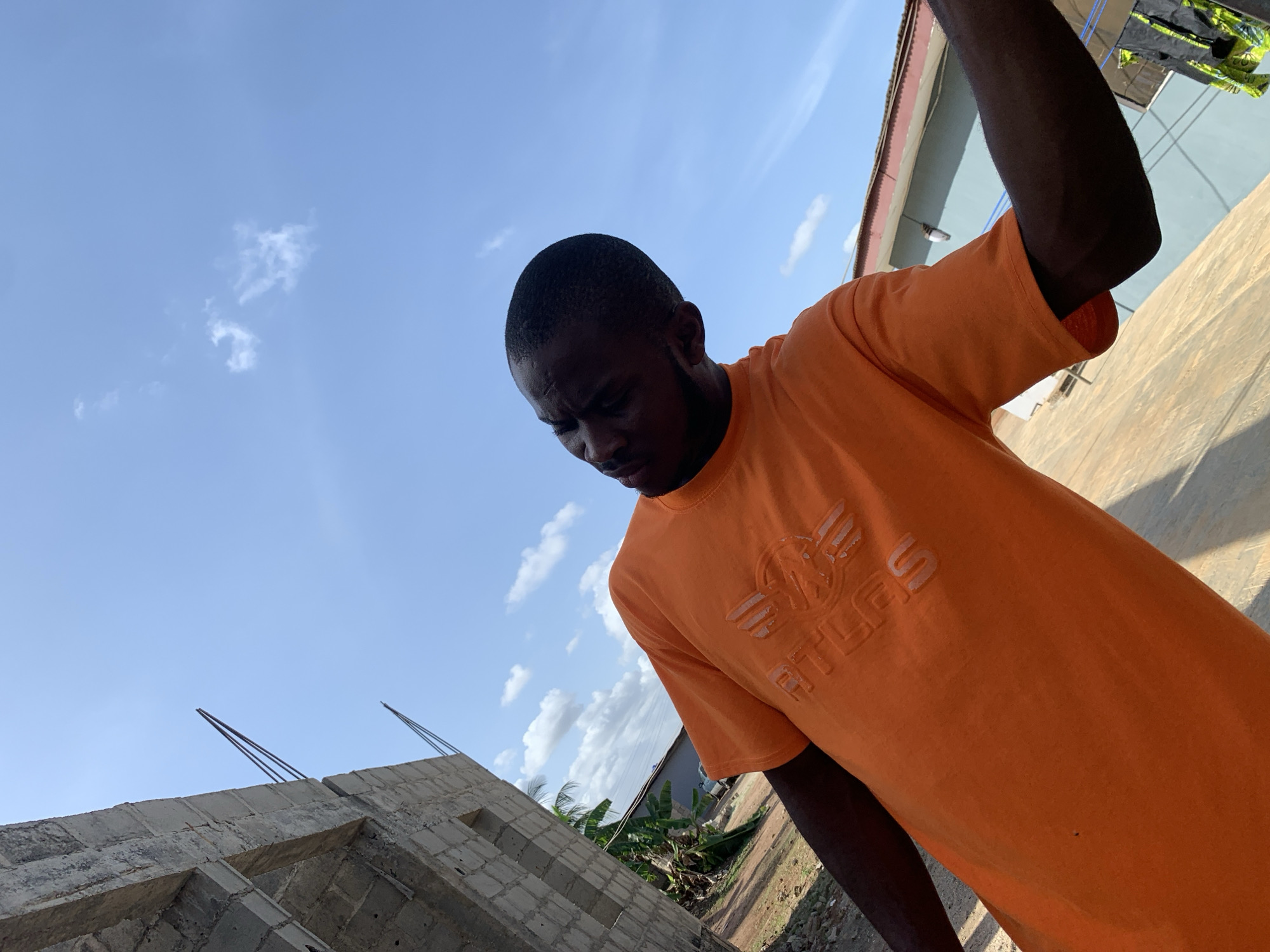 The image shows a man in an orange t-shirt with the word "ATLAS" on it, standing next to a partially constructed building of grey blocks, with metal rebar protruding from the top. The background includes a blue sky with scattered white clouds. In the distance are trees and a building. The location is Ikorodu, Nigeria. The man is looking downwards.