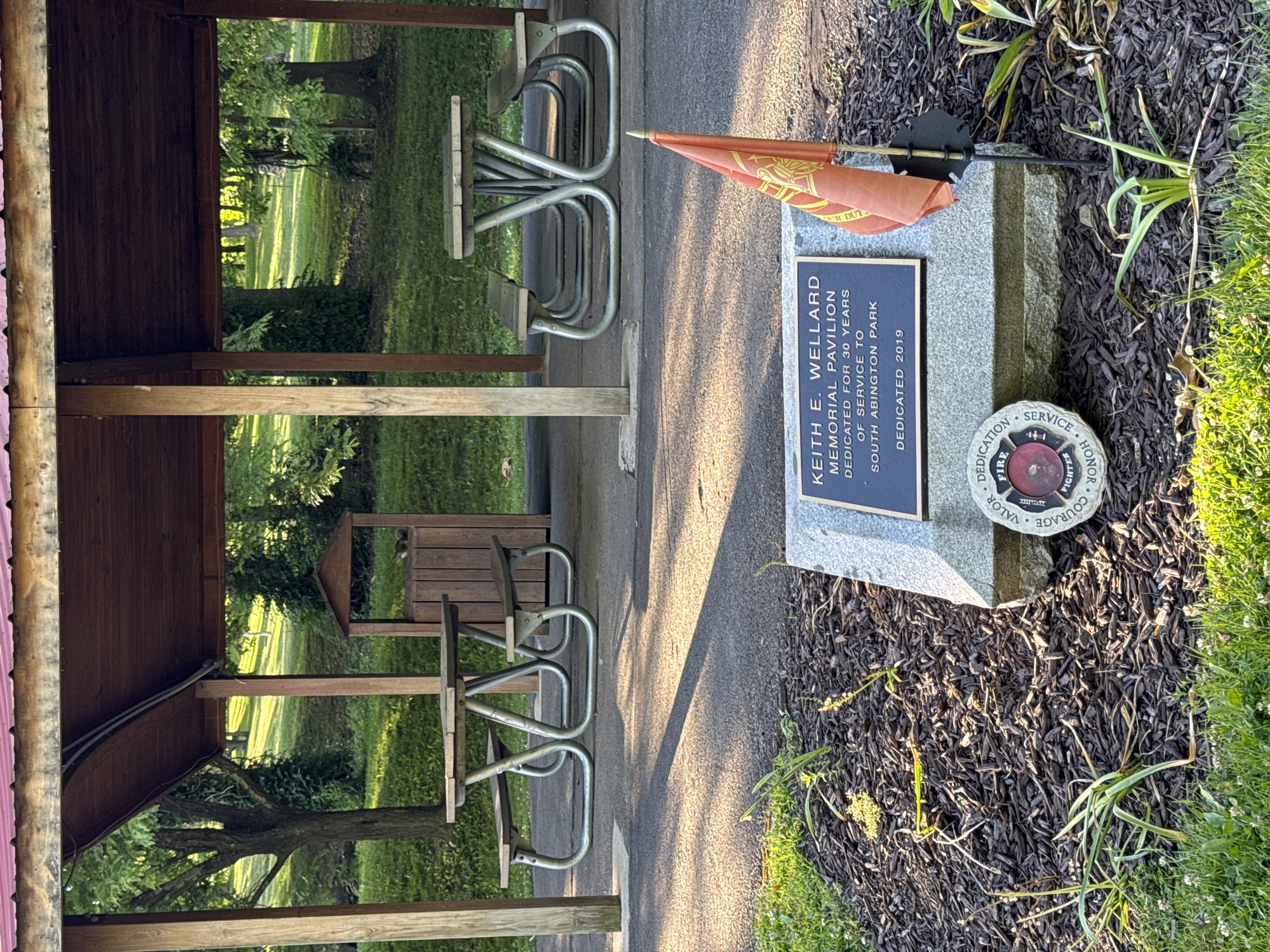 The image shows a memorial pavilion and picnic tables in a park setting, likely South Abington Park in South Abington Township, United States of America. In the foreground, a stone monument with a bronze plaque reads "KEITH E. WELLARD MEMORIAL PAVILION DEDICATED FOR 30 YEARS OF SERVICE TO SOUTH ABINGTON PARK DEDICATED 2019". A small orange flag with a crest is planted in front of the monument. To the left of the monument is a circular stone emblem with the words "VALOR COURAGE HONOR SERVICE FIRE FIGHTER DEDICATION". Behind the monument, several picnic tables with benches are visible under a wooden pavilion with a reddish-brown roof. The ground is paved with asphalt and covered with mulch in the memorial area. Trees and grassy slopes form the background, with sunlight filtering through the leaves.