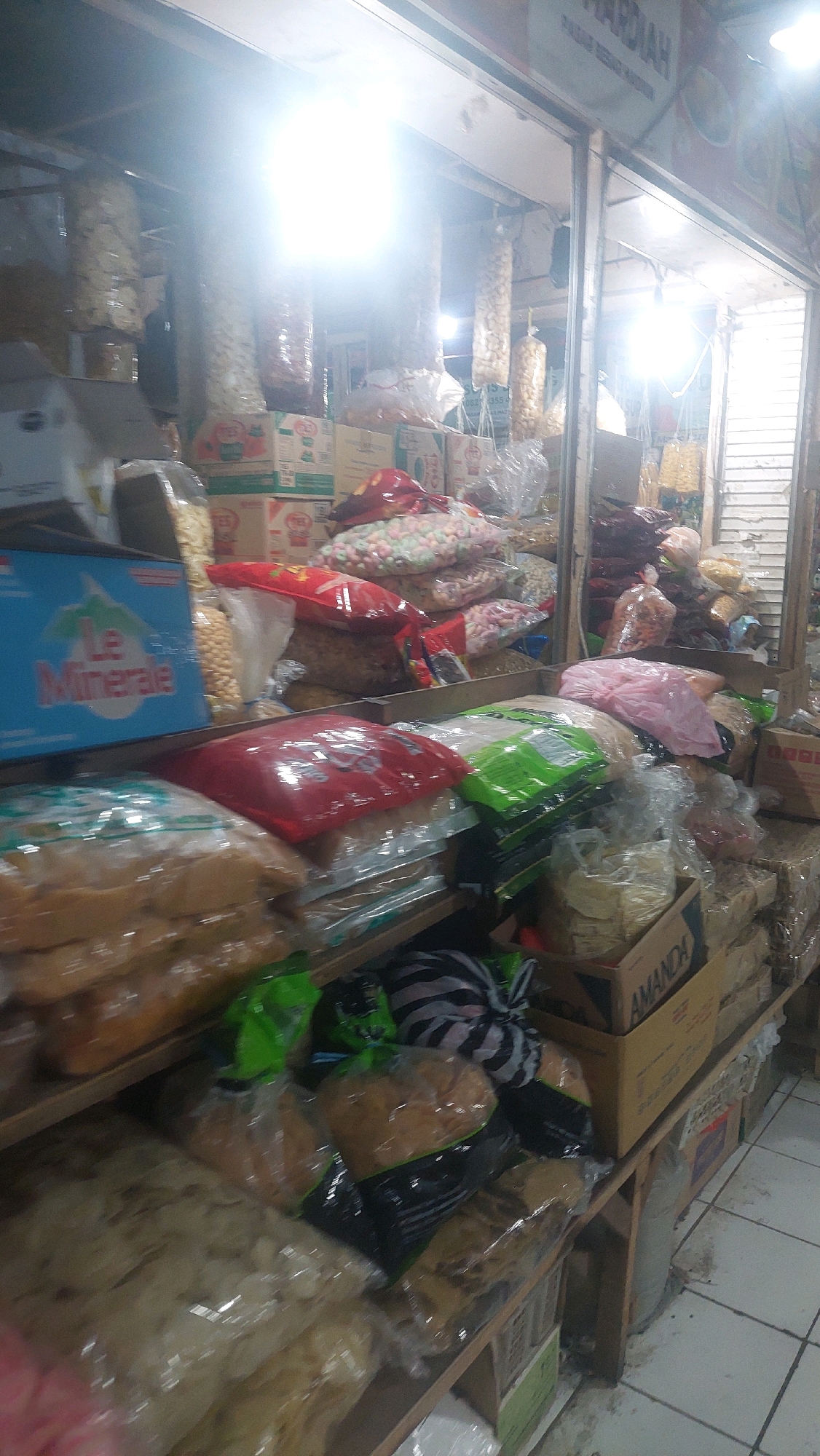 The image shows a stall in a market in Madiun, Indonesia, filled with various packaged food items. The stall is dimly lit, with bright overhead lights illuminating the products. Shelves are packed with bags and boxes of snacks, including chips, crackers, and candies, some of which are transparently packaged, allowing the contents to be visible. Several large plastic bags filled with snacks hang from the ceiling. Boxes are stacked on shelves and on the floor. The overall impression is of a well-stocked food vendor's stall.