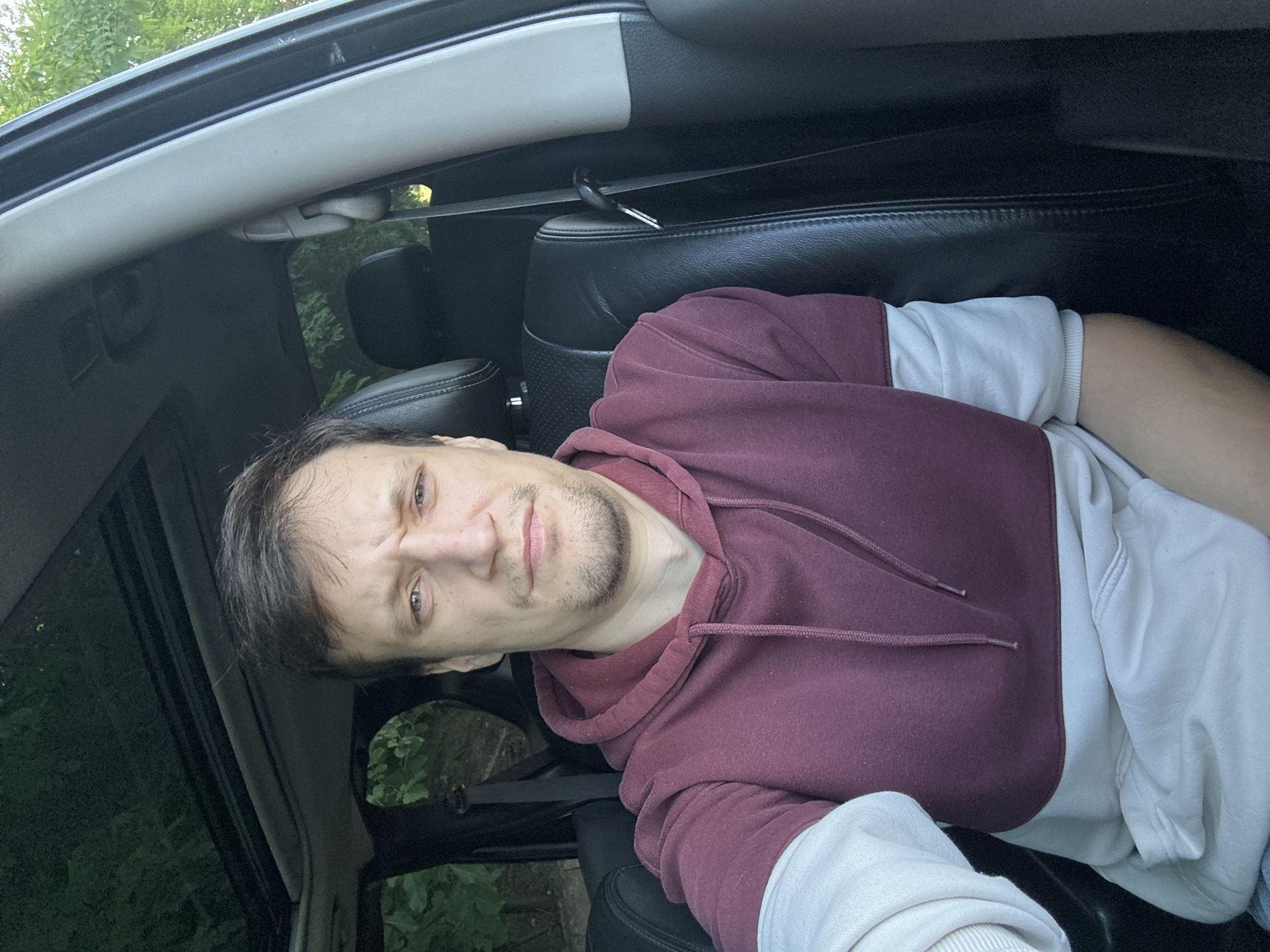 A Caucasian male with short brown hair and a light beard is sitting in the driver's seat of a car, facing the camera. He is wearing a maroon and light gray hoodie. The car has a sunroof, which is open, and dark leather seats. The background shows greenery and trees, suggesting an outdoor location. The image is a selfie, taken from a slightly low angle. The man's facial expression is neutral. The exact location in Makiv, Ukraine is not discernible from the visual information in the image.