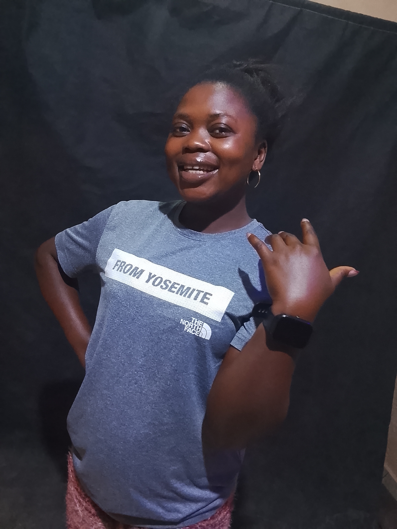 A young, dark-skinned woman is posing in front of a dark, possibly draped, background. She is wearing a grey t-shirt with "FROM YOSEMITE THE NORTH FACE" printed on it, and a digital watch on her left wrist. Her right hand is raised in a "hang loose" or "shaka" gesture. She has earrings and is smiling. The woman's location is Unknown, Unknown.