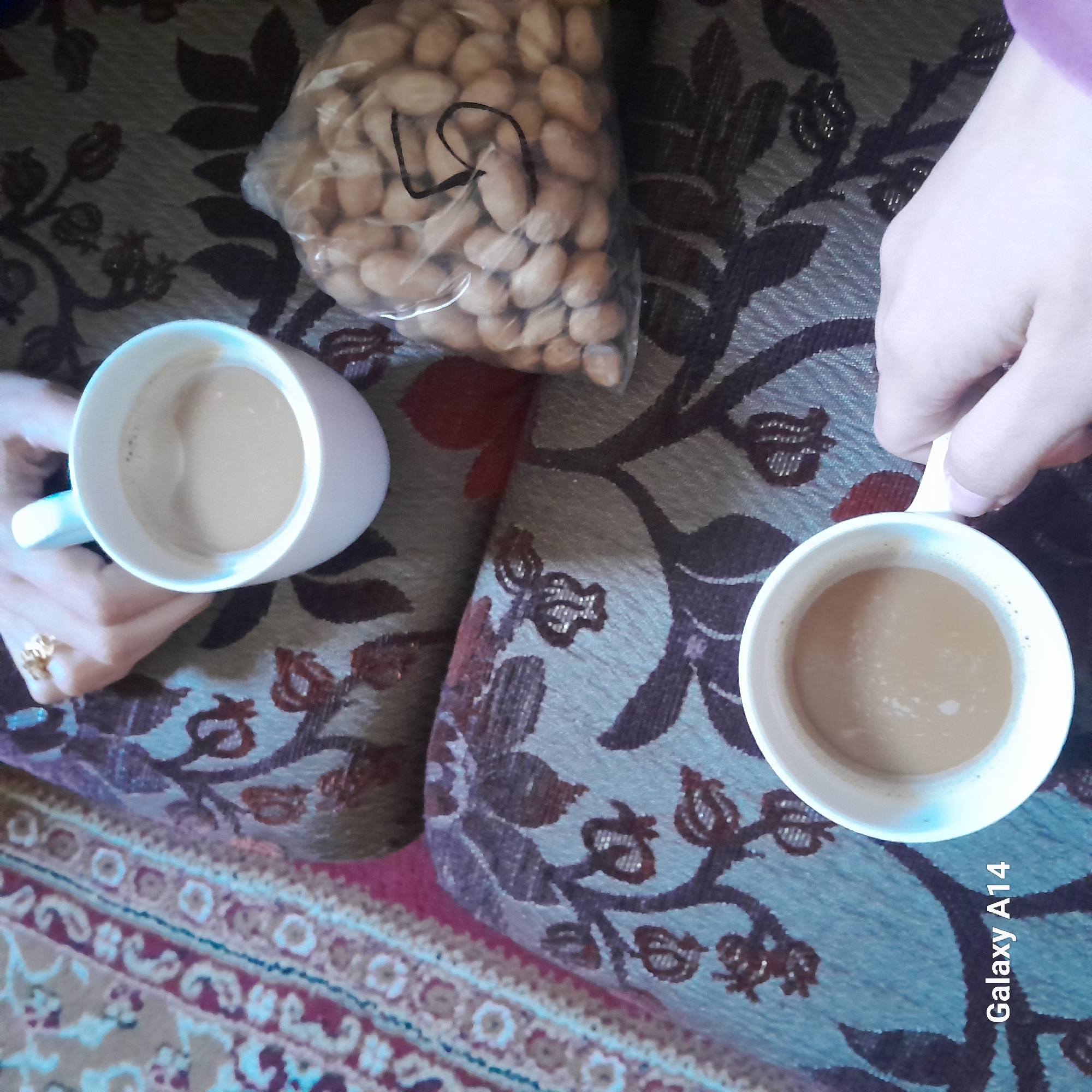 The image shows two people having coffee together, with a bag of nuts between them. The scene is indoors, possibly in a home, given the patterned couch and rug. One person's hand is visible, holding a white mug of coffee, while the other person's hand is holding the other mug. A gold ring is visible on one person's hand. The bag of nuts is a clear plastic bag filled with light-colored nuts. The location is unknown.