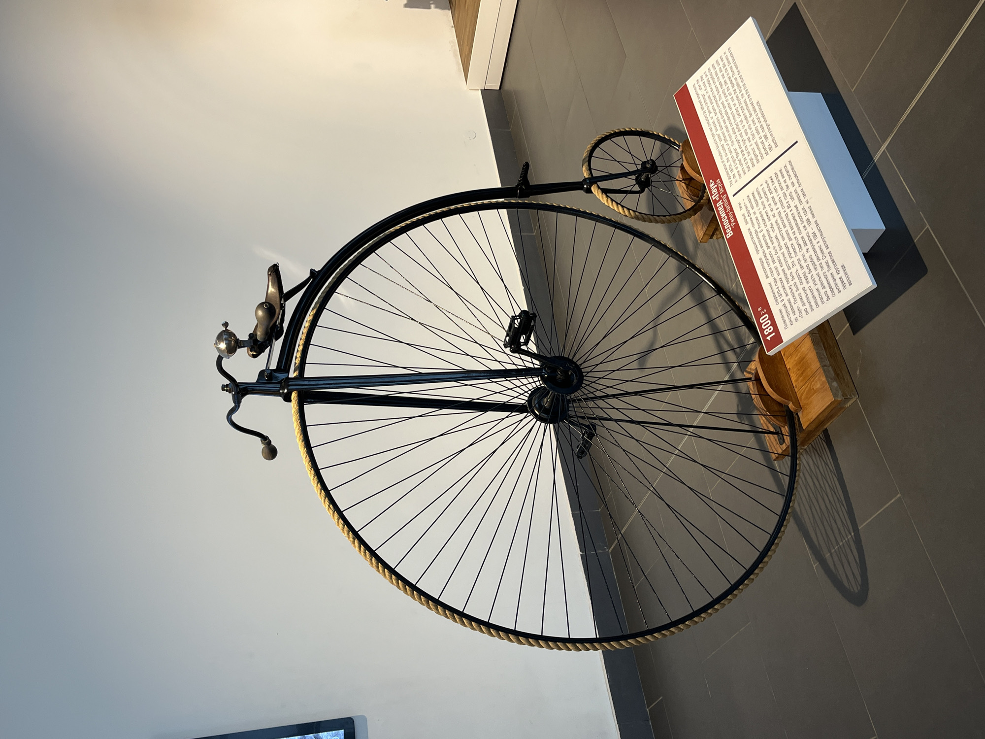 This media file shows a vintage "penny-farthing" bicycle displayed in a museum. The bicycle, also referred to as a "spider" bicycle in Russian, features a prominently large front wheel and a much smaller rear wheel. It is black with a rope-like material around the rim of the front wheel and has a bell and handlebars at the front. The bicycle is mounted on a wooden stand, casting a shadow on the tiled floor. To the right of the bicycle is a display board with text. The text, written in both Russian and English, describes the history of the penny-farthing bicycle, noting its popularity in the 1870s and its characteristics, including its dangerous nature due to a high center of gravity and the inconvenience of mounting and dismounting. It also mentions the notable 1884-1886 round-the-world bicycle trip by Englishman Thomas Stevens on such a bicycle. The background consists of a plain white wall with a subtle shadow. The lighting suggests an indoor setting, possibly a museum or exhibition space. The overall scene is static, focusing on the historical artifact and its accompanying information.