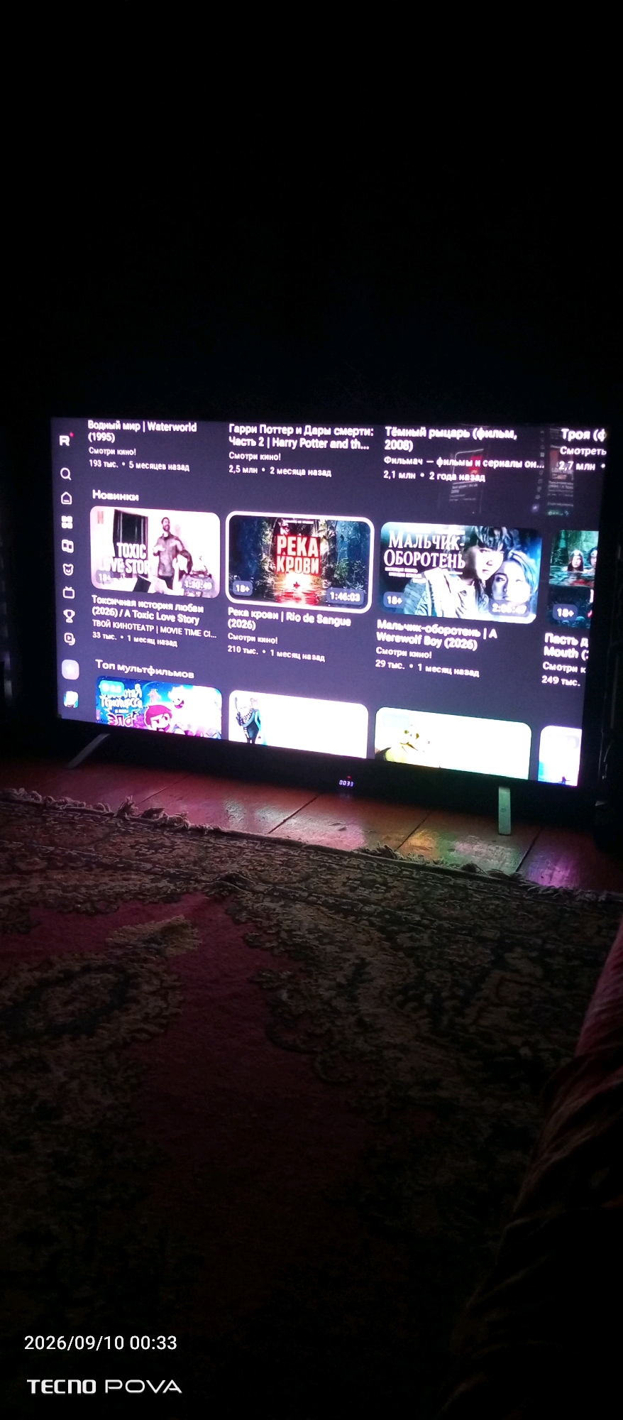 A television screen displays a collection of movie and series thumbnails. The screen is dark, with the content glowing. The layout suggests a streaming service or video library interface. Top entries include "Waterworld," "Harry Potter and the Deathly Hallows - Part 2," and "Teen Wolf." Below these are sections for "New Releases" and "Top Animated Films."
In the "New Releases" section, specific titles visible are "Toxic Love Story (2026)," "Rio de Sangue (2026)," and "Werewolf Boy (2026)." These thumbnails are accompanied by viewing statistics like "193 тыс.," "2.5 млн," "210 тыс.," "29 тыс.," and release time information such as "5 месяцев назад," "2 месяца назад," "1 месяц назад," and "2 года назад."
The screen is positioned in a dimly lit room, with a patterned rug visible on the floor in the foreground. The rug appears to be made of a coarse material and has a reddish-brown hue. The overall environment suggests a home entertainment setting. The lower portion of the image shows a timestamp "2026/09/10 00:33" and the text "TECNO POVA." The location of Kudeyevskiy, Russia, is not directly identifiable from the visual elements of the image itself.