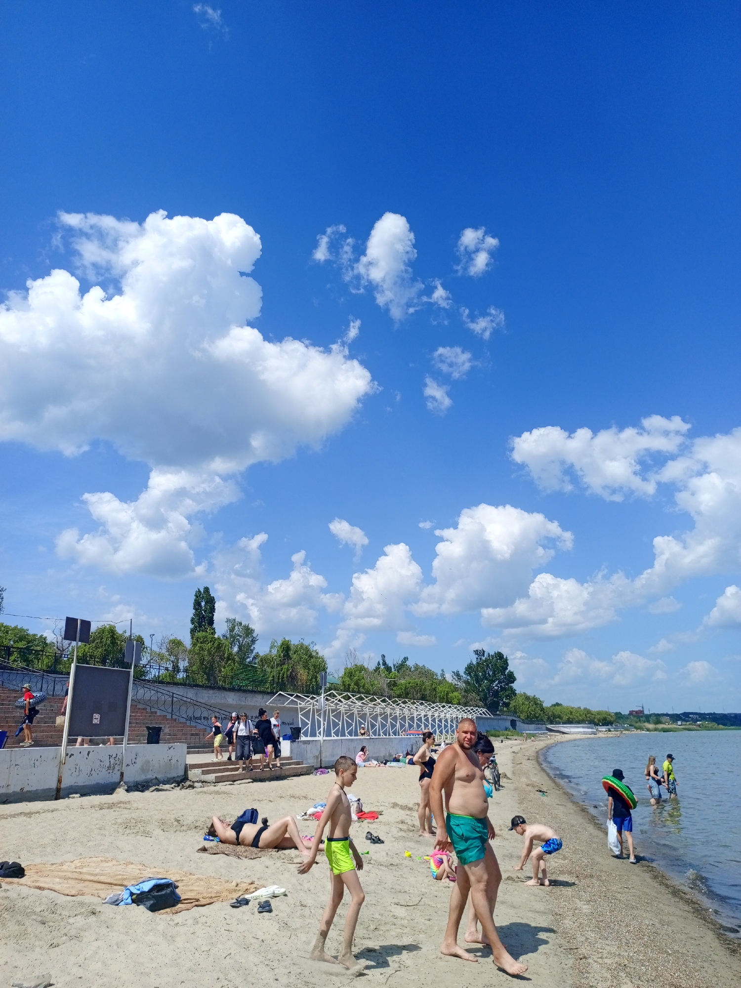 The image depicts a beach scene on a sunny day with a bright blue sky dotted with white clouds. A crowd of people are enjoying the beach: some are sunbathing on towels, others are walking along the shore, and a few are in the water.
A man wearing green shorts is standing on the sand in the foreground. A child in bright green shorts is walking nearby. Further down the beach, people are playing in the sand and wading in the water.
In the background, there is a low wall, a row of trees and some structures that resemble shelters or covered walkways. The location is likely a city beach, possibly in an urban area. The city is unknown.