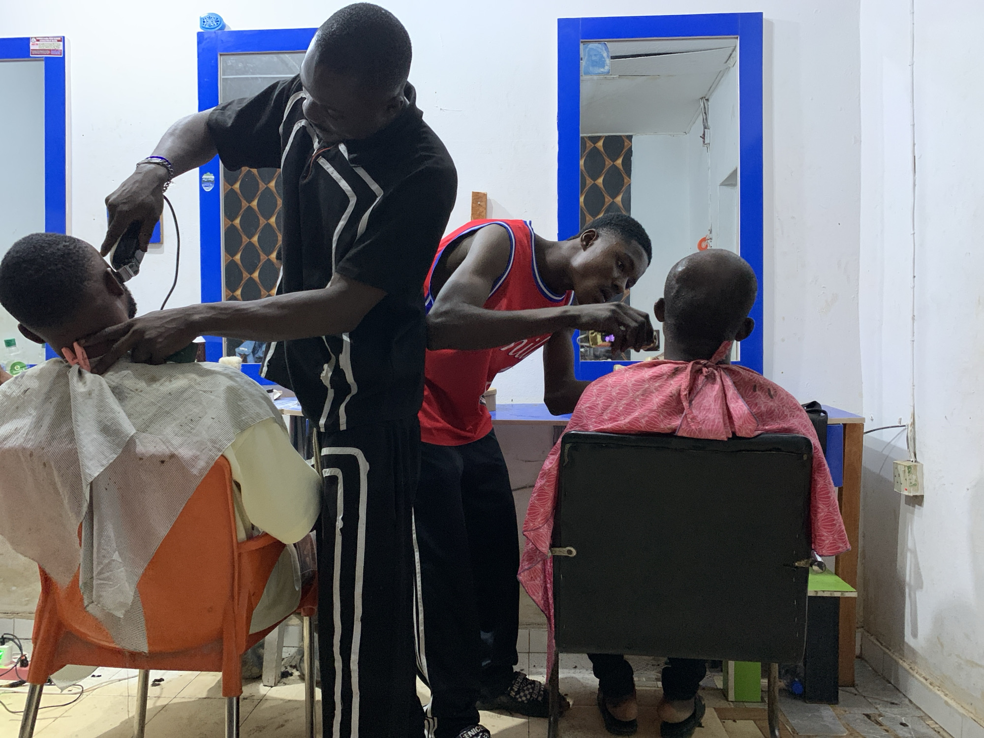 In a simple barbershop, likely in Gbeleko, Nigeria, two barbers give haircuts to their customers. One barber, wearing a black tracksuit with white stripes, uses an electric clipper on a seated customer draped in a white and gray barber cape, seated on an orange plastic chair. The second barber, clad in a red basketball jersey, focuses intently on the back of the head of a customer in a black office chair covered in a pink and red cape. Blue-framed mirrors are mounted on the white walls. The floor shows signs of wear and is speckled with debris. An electrical outlet is visible on the wall.
