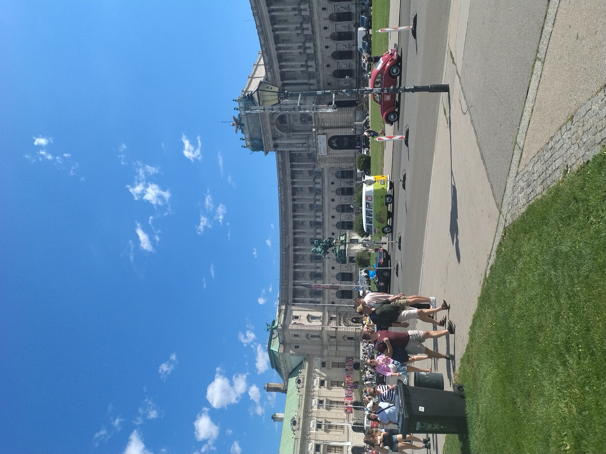 The image shows a daytime outdoor scene in front of a large, ornate building in Vienna, Austria, likely the Hofburg Palace complex, characterized by its grand architecture and numerous columns. The sky is bright blue with scattered white clouds. In the foreground, several people are walking on a paved walkway next to a grassy area. To the left, a large black trash bin is visible.
Further back, several vehicles are present on a street. A distinctive vintage green and white camper van is parked on the grass, and a classic red Volkswagen Beetle is driving on the street. The building itself has a light-colored facade and a green roof, with statues and decorative elements visible along its upper portions. There are flags or banners hanging from poles in front of the building. The overall impression is of a sunny day with tourists and locals enjoying the public space and historical architecture.