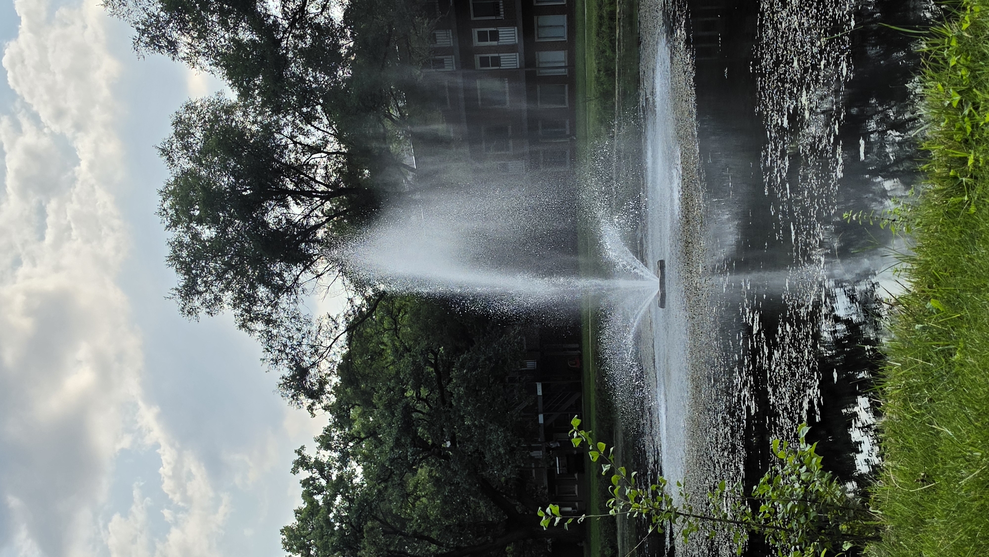 A fountain sprays water in a pond in front of a brick apartment building with white trim windows. The pond has lily pads and reflections of the trees and building. Green grass and plants are in the foreground, and lush green trees are behind the pond and building. The sky is partly cloudy with a mix of white and grey clouds.