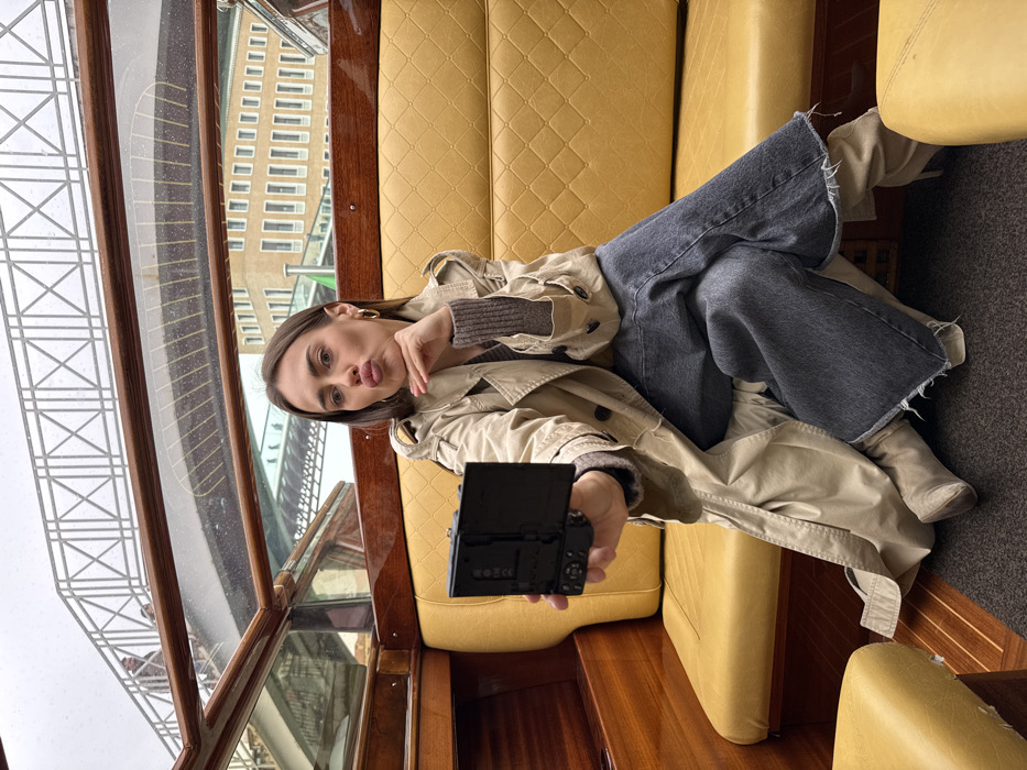 A young adult female is depicted seated inside a water taxi on a canal in Venice, Italy. She wears a light beige trench coat, a brown ribbed sweater, dark grey wide-leg jeans with frayed hems, and light beige high-heeled boots. The subject holds a black compact camera with a flip-out screen in her right hand, aimed at herself, and has her left hand positioned under her chin while her lips are pursed. The boat's interior features polished wooden paneling and mustard yellow diamond-quilted seating. Through the boat's windshield, an overcast sky is visible above a grey metal pedestrian bridge with a lattice-patterned railing, under which the boat is positioned. Indistinct figures are present on the bridge. A multi-story building with vertical windows stands in the background to the right.