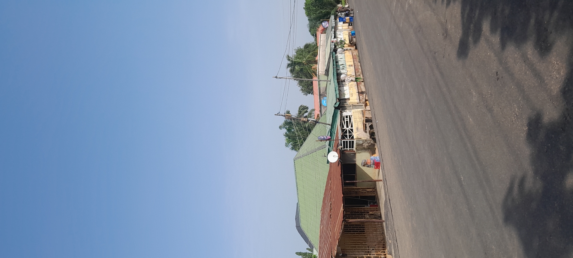 The image depicts a row of small, weathered shops along a paved road in Jalingo, Nigeria. The buildings have a mixture of corrugated metal and tiled roofs, with the most prominent structure featuring a bright green, multi-faceted roof. Many of the shops have open fronts, showcasing shelves with items, and some have barred or caged entrances. A satellite dish is visible on one of the buildings. In front of the shops, crates of bottled drinks are stacked near the road. A few people are visible in the distance walking away from the camera, some carrying bags. Trees, including palm trees, are present behind the buildings. Utility poles with wires run along the side of the road, indicating infrastructure. The sky is clear and blue, suggesting a sunny day. The overall scene suggests a commercial area in a Nigerian city.