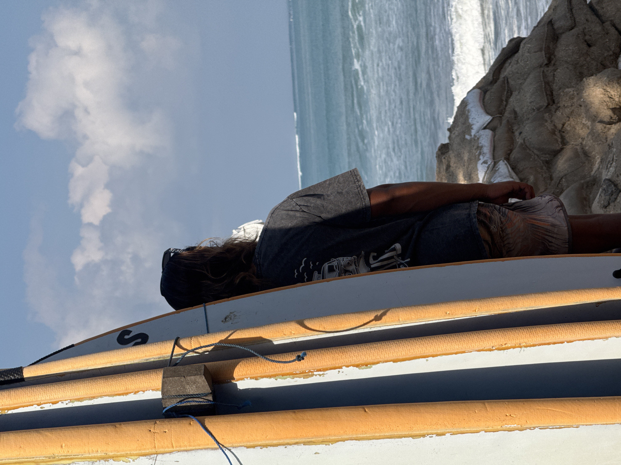 The image captures a person standing next to several surfboards on a beach in Kuta, Indonesia. The surfboards are leaning against each other, with their orange foam edges facing forward. A black letter "S" is visible on one of the surfboards. The person, viewed from behind, is wearing a dark t-shirt and patterned shorts, with sunglasses perched on their head. They are looking out towards the ocean, where waves are breaking on the shore. In the foreground, a rocky outcrop, reinforced with sandbags, is visible. The sky above is blue with scattered white clouds, and the sun is casting shadows on the surfboards and the ground.