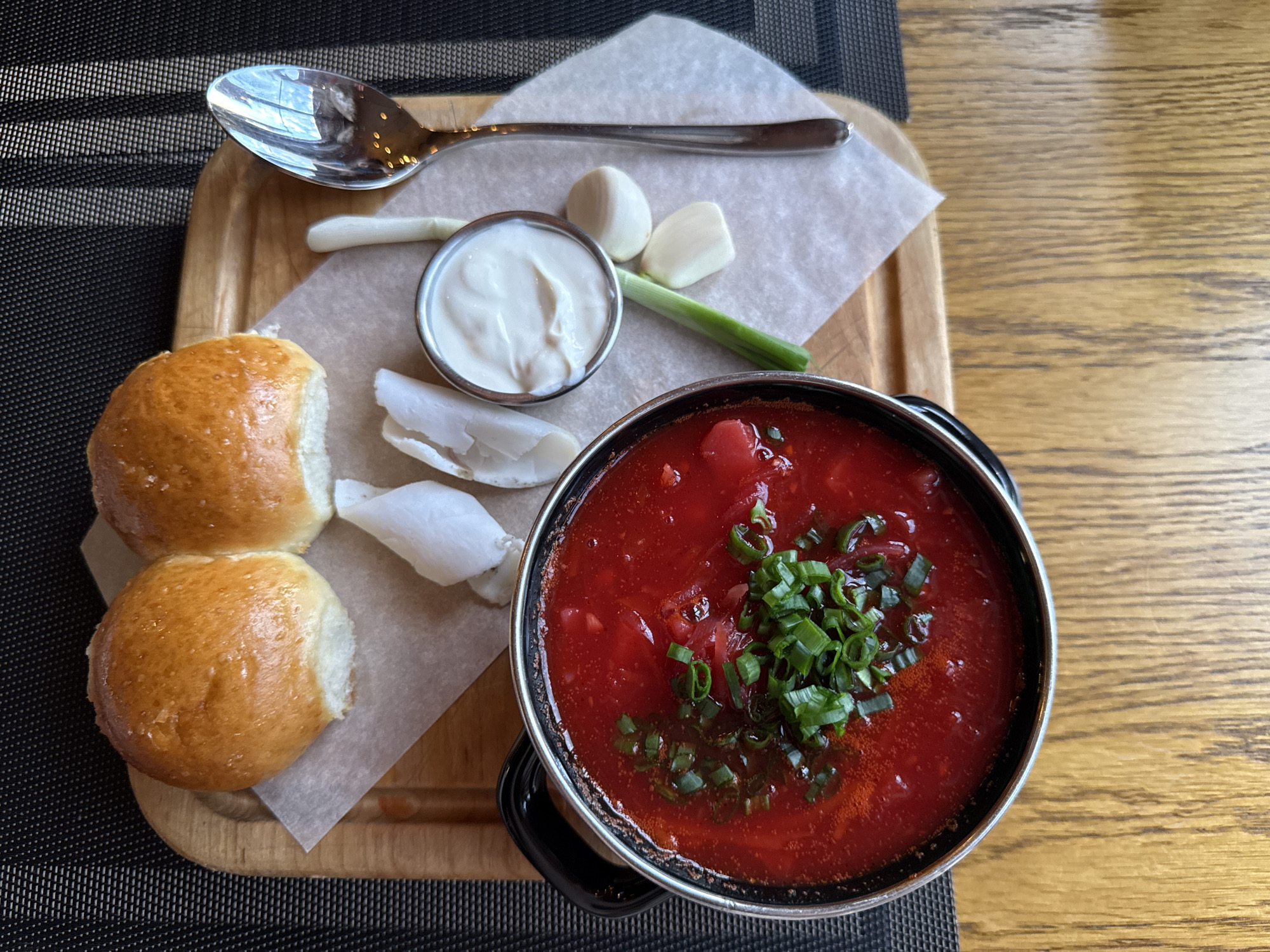 A top-down, eye-level shot shows a rustic wooden table in Olginka, Russia, set with a traditional meal. In the foreground, a black metal pot of vibrant red borscht, garnished with chopped green onions, is the centerpiece. To its right, a small silver bowl holds creamy white sour cream, accompanied by two cloves of garlic and a green onion stalk. Behind the soup, two golden-brown, fluffy buns are nestled on a piece of parchment paper. A gleaming silver spoon rests to the right of the sour cream. The background is a textured, dark surface, possibly a placemat, over the warm tones of the wooden table. The lighting suggests an indoor setting, possibly a restaurant.