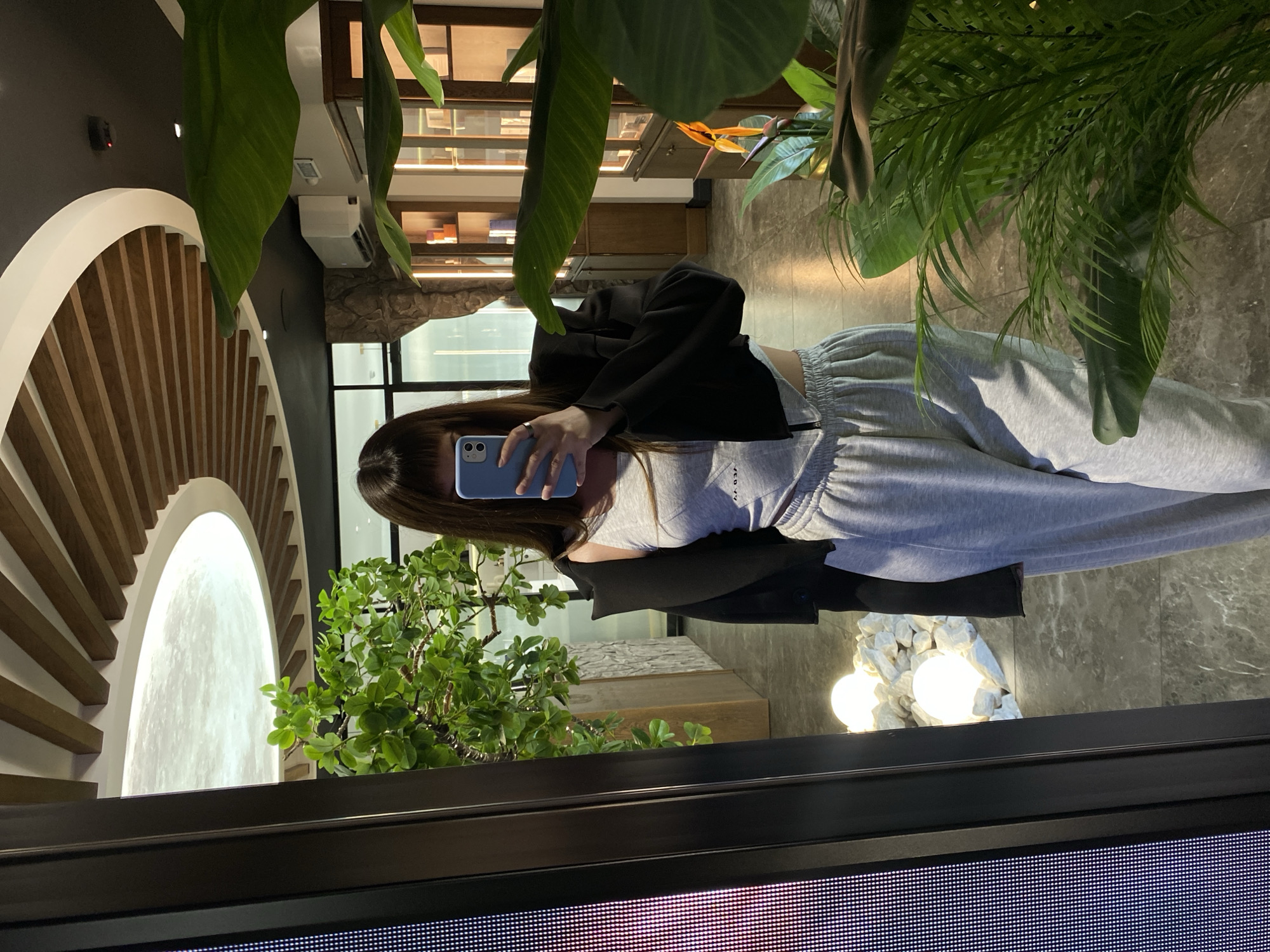 The image shows a person taking a mirror selfie in what appears to be an indoor public space, possibly a commercial establishment like a store or a cafe. The person is wearing a gray crop top and gray sweatpants, with a black jacket draped over their shoulders. They are holding a blue smartphone in their right hand, positioned to capture the reflection.
The environment is decorated with plants, including a large potted plant in the foreground to the right, and a smaller tree-like plant to the left. In the background, there are shelves stocked with various items, suggesting retail display. The ceiling features a modern design with curved wooden elements and a circular illuminated panel. There are also decorative lights placed amongst white stones on the floor, visible in the mirror's reflection. The flooring appears to be tiled.
There is no information in the image that allows for the identification of the city as Votkinsk, Russia, or any specific location within that city. The description is limited to the visual elements present in the photograph.