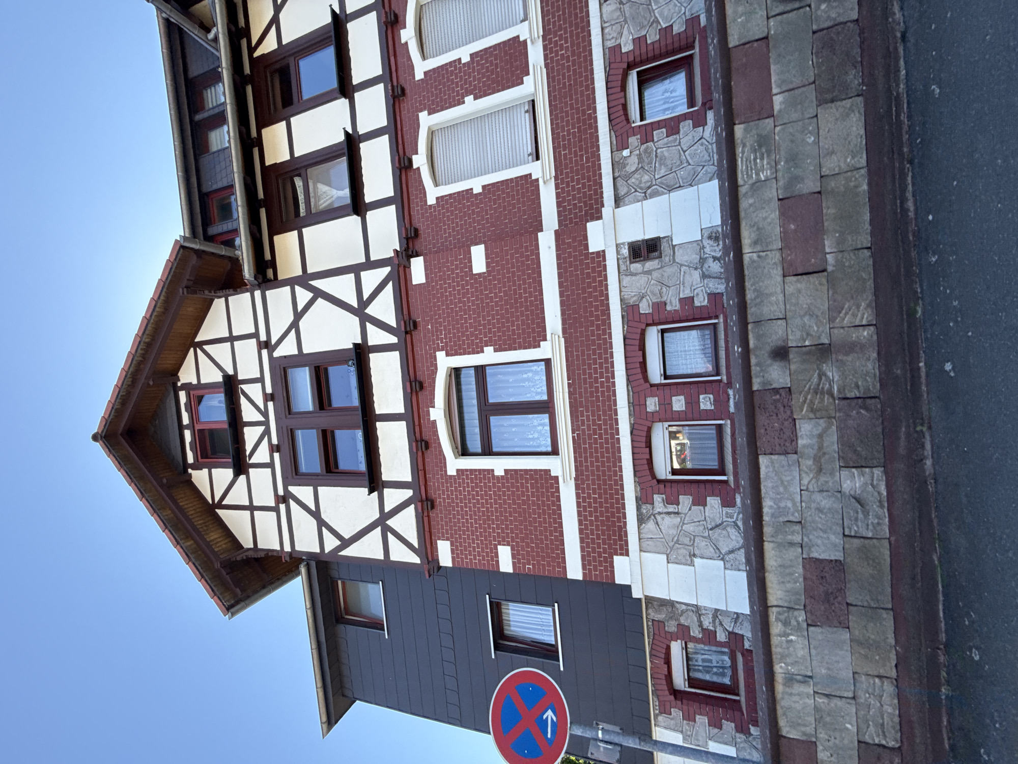 The image shows a view of the exterior of a building in Bad Sachsa, Germany. The building features a distinctive timber-framed upper section with white infill panels and dark brown timbers, indicative of a traditional German architectural style. Below this, the building has a red brick facade with white decorative elements. The lower sections are clad in stone. There are multiple windows visible, some with white curtains and others with closed shutters. In the foreground, a stone wall separates the building from the street. A traffic sign indicating a no-parking zone with an arrow pointing to the right is visible on the left side of the image. The sky is clear and blue.
