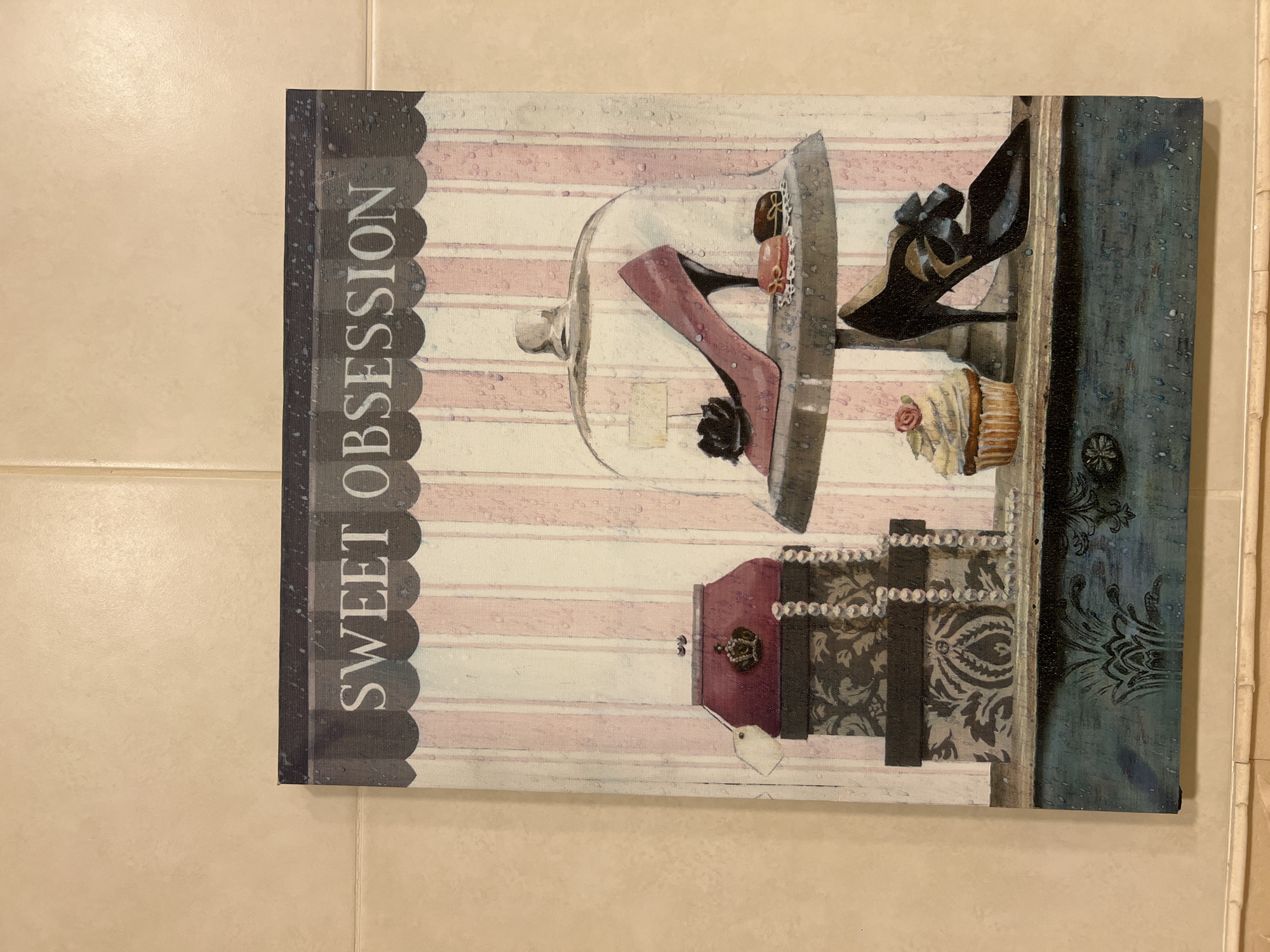 The image shows a piece of wall art with the words "SWEET OBSESSION" printed across the top. The artwork depicts a still life of a pink high-heeled shoe under a glass dome, a cupcake, a black high-heeled shoe with a bow, and several gift boxes with pearls draped over them. The background features pink and white stripes, and the overall effect is a whimsical, feminine aesthetic. The artwork is hanging on a tiled wall, the tiles being a neutral, beige color. The lighting appears to be from above and slightly to the left. No people are present in the image. The image is a photograph of the artwork and its surrounding wall. The wall art is positioned centrally on the wall, approximately at eye level.