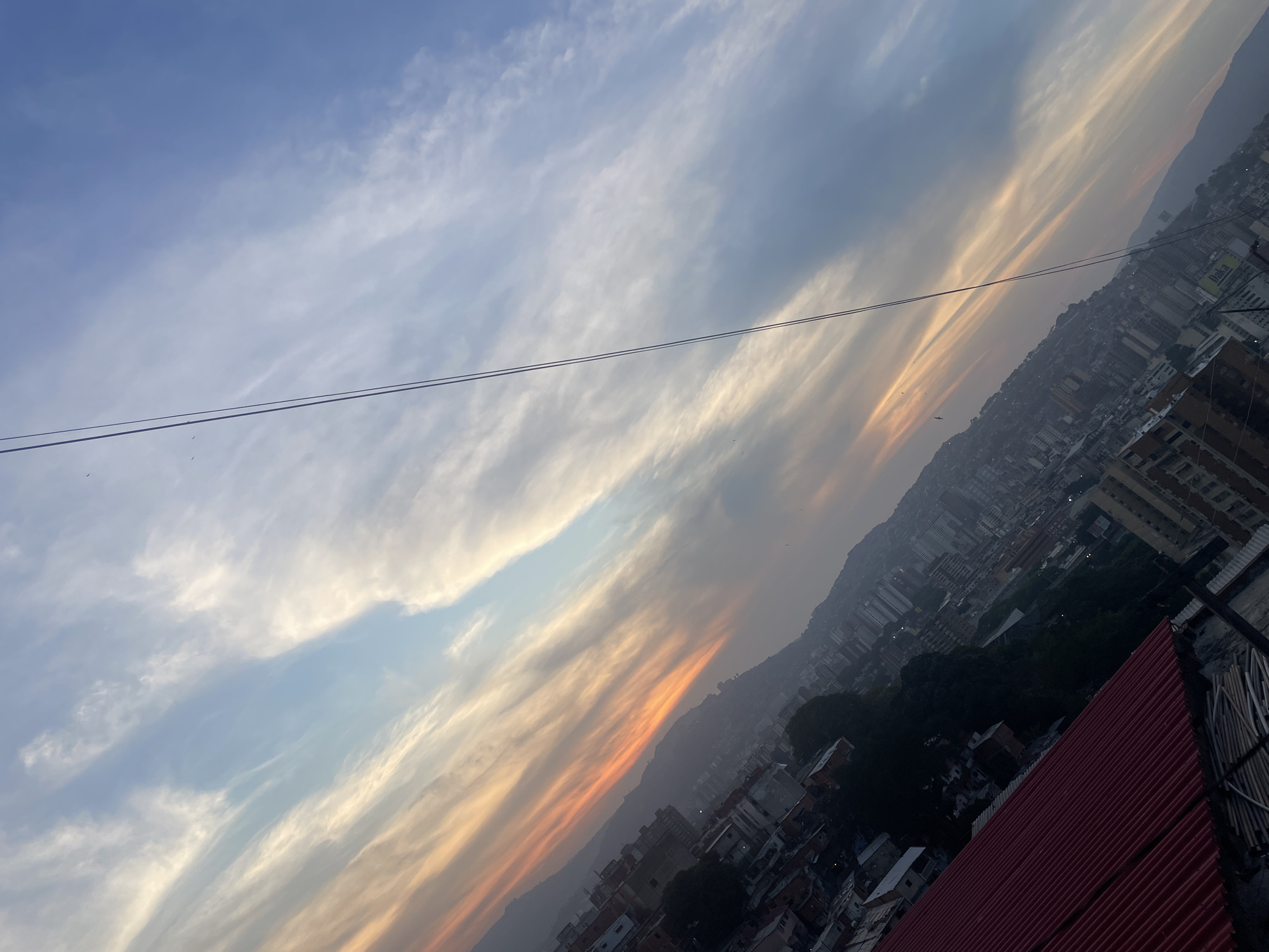 The image shows a view of Caracas, Venezuela at dusk. The sky is streaked with clouds, transitioning from blue to orange hues. A pair of overhead wires stretch across the top of the frame. Below the sky, a densely populated area with buildings of various sizes and architectural styles cascades down a hillside. The overall impression is of a city nestled within a mountainous landscape during a colorful sunset.