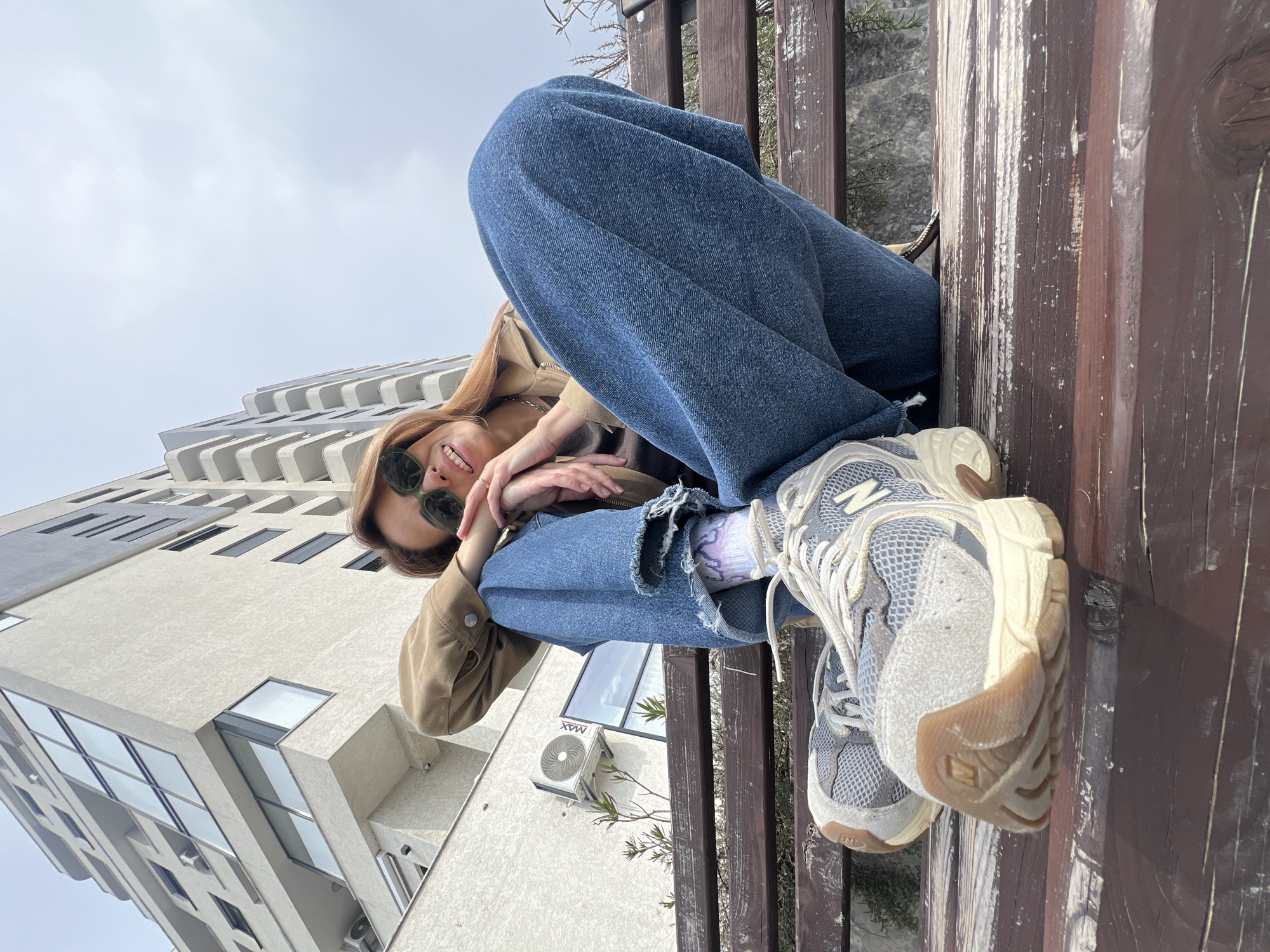 An outdoor, low-angle photograph captures a female subject seated on a weathered, dark brown wooden slatted bench in Bar, Montenegro, under an overcast sky. The subject is depicted from the waist up, smiling towards the camera while wearing dark green sunglasses. She is dressed in a light brown jacket over a brown top, wide-leg blue jeans with frayed hems, white socks featuring a purple tie-dye pattern, and grey and beige New Balance sneakers. Her legs are bent, with her forearms resting on her knees; the right sneaker is prominently visible in the foreground.
Behind the subject, a low-growing green shrubbery borders the base of a modern, multi-story building. This building is light-colored with textured stucco, characterized by angular architectural elements and large rectangular windows. An outdoor air conditioning unit, branded "MAX," is mounted on its lower exterior wall. Further in the background, another multi-story structure with uniform white balconies is visible against the light grey sky. The scene is lit by diffused natural light, indicating an absence of direct sunlight.